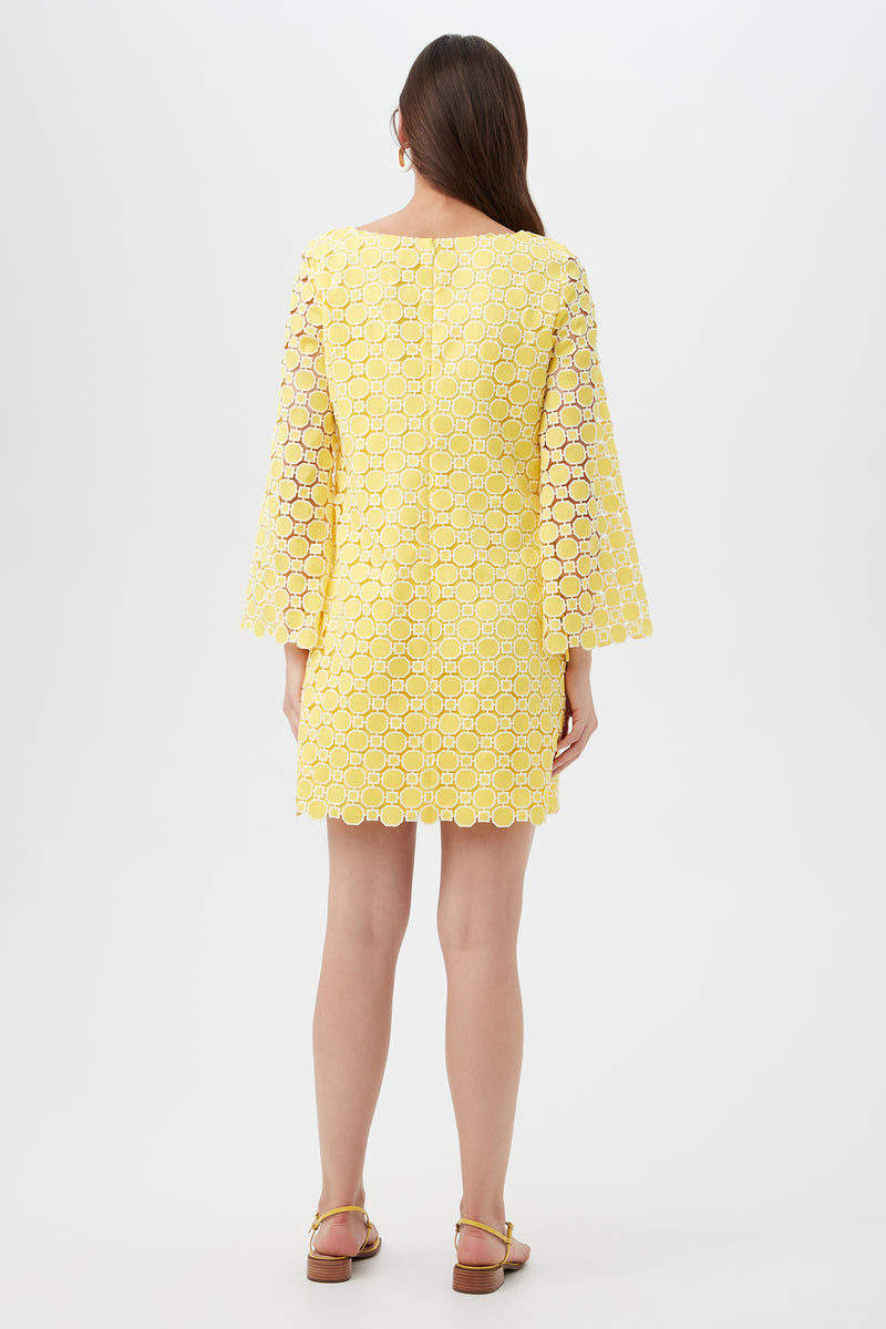 A woman faces away, wearing the Ariella 2 Long-Sleeve Dress—a knee-length yellow dress with circular lace pattern and wide sleeves. She pairs it with sandals, her straight brown hair down, standing against a plain light background.