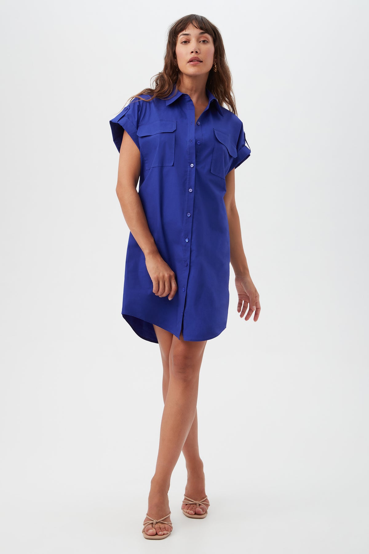A woman poses in the Bonsoir Short-Sleeve Shirt Dress in Cobalt Cove, a loose-fitting, button-down style with two chest pockets. She stands against a plain white background, wearing strappy sandals and her long brown hair down.