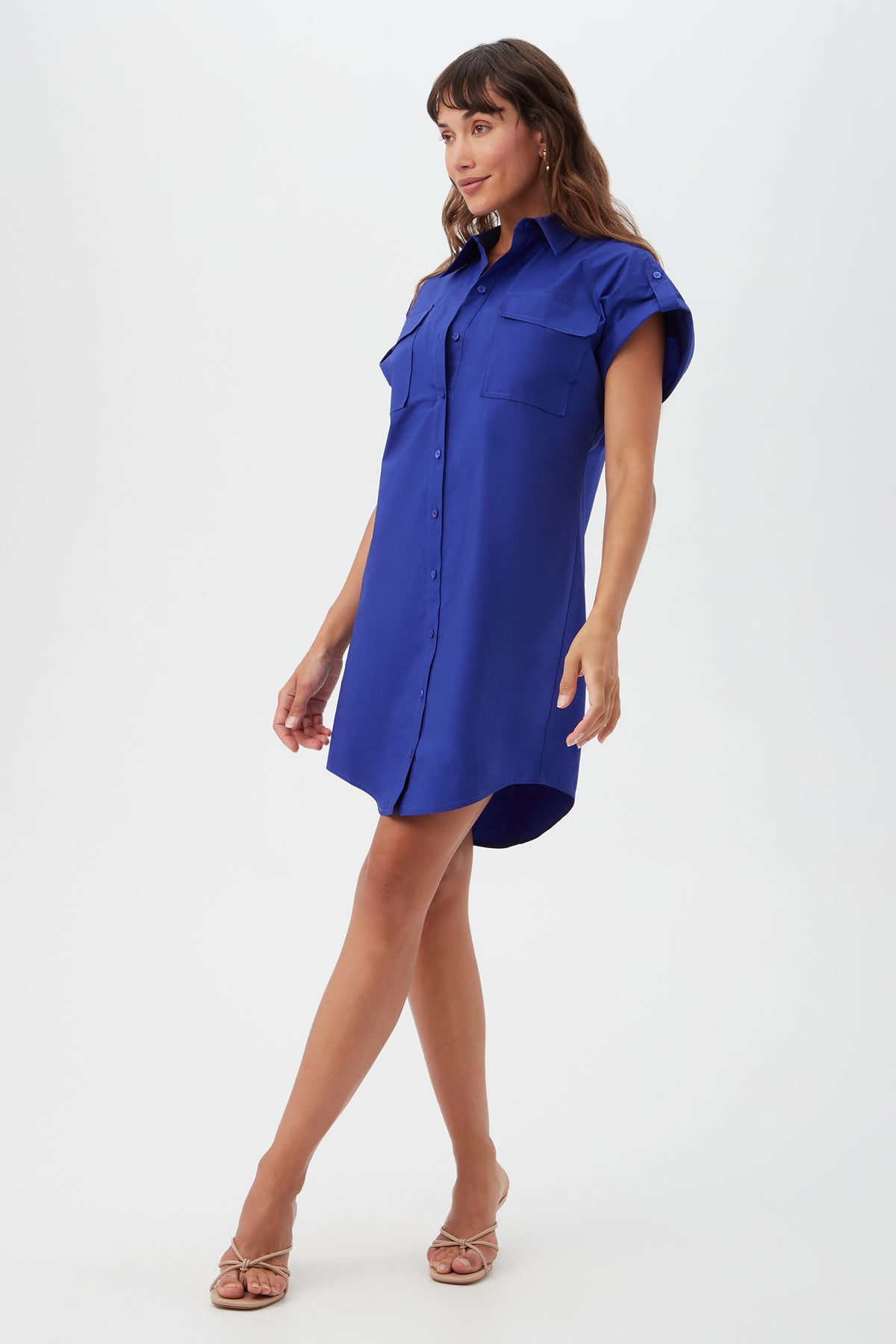 A woman poses against a white background, wearing the Bonsoir Short-Sleeve Shirt Dress in bright blue with button-up front and two chest pockets, paired with beige heeled sandals.