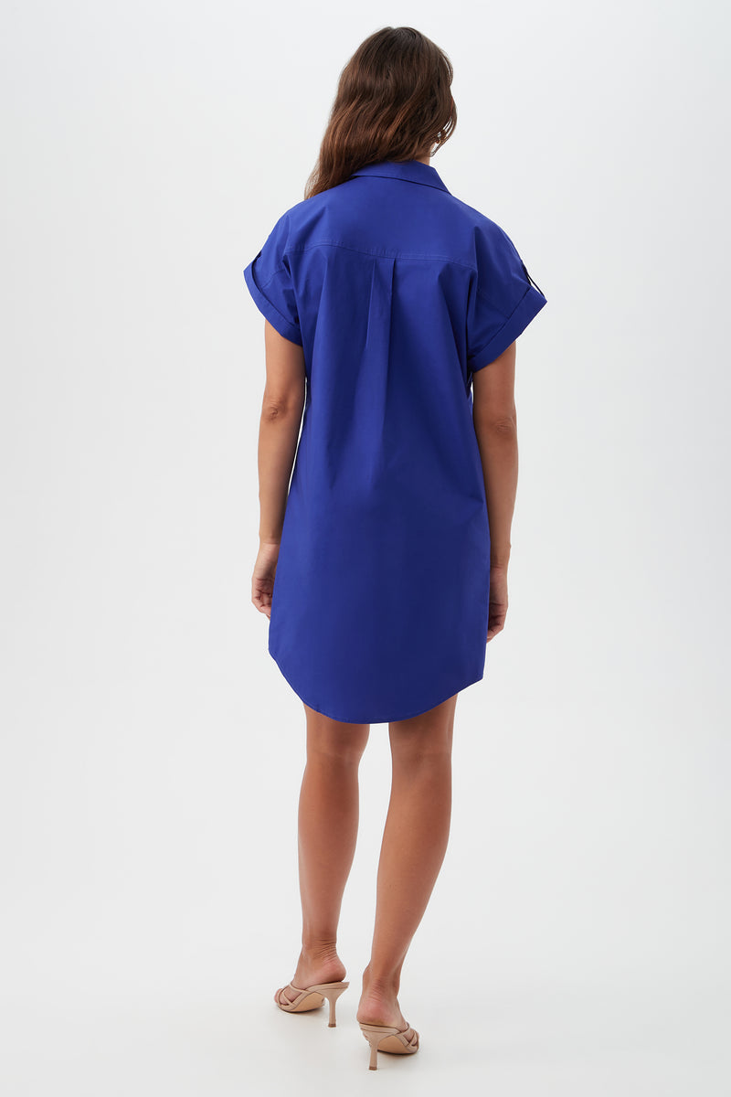 A woman with long wavy brown hair wears the Bonsoir Short-Sleeve Shirt Dress in royal blue, knee-length and loose-fitting, paired with beige heeled sandals. She stands facing away against a plain white background.