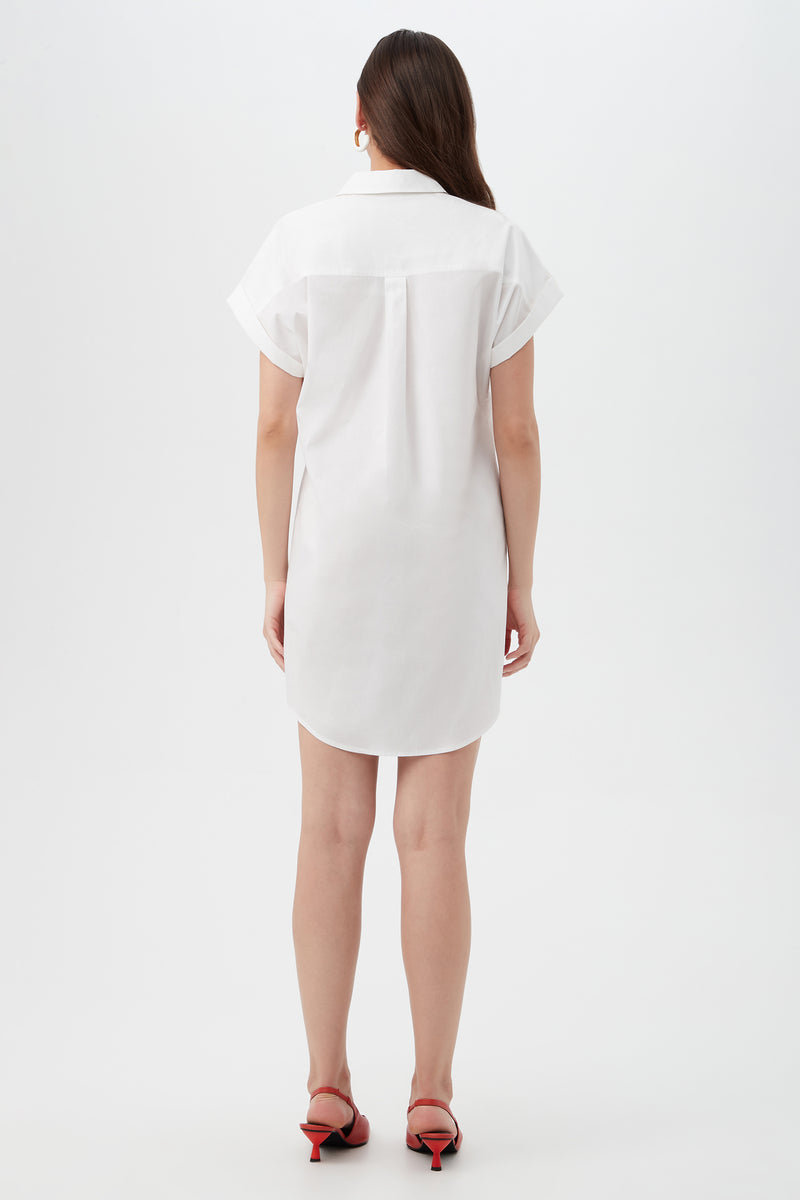 A woman with long brown hair stands facing away in the Bonsoir Short-Sleeve Shirt Dress, a short white dress with cuffed sleeves, paired with red heeled sandals against a plain white background.