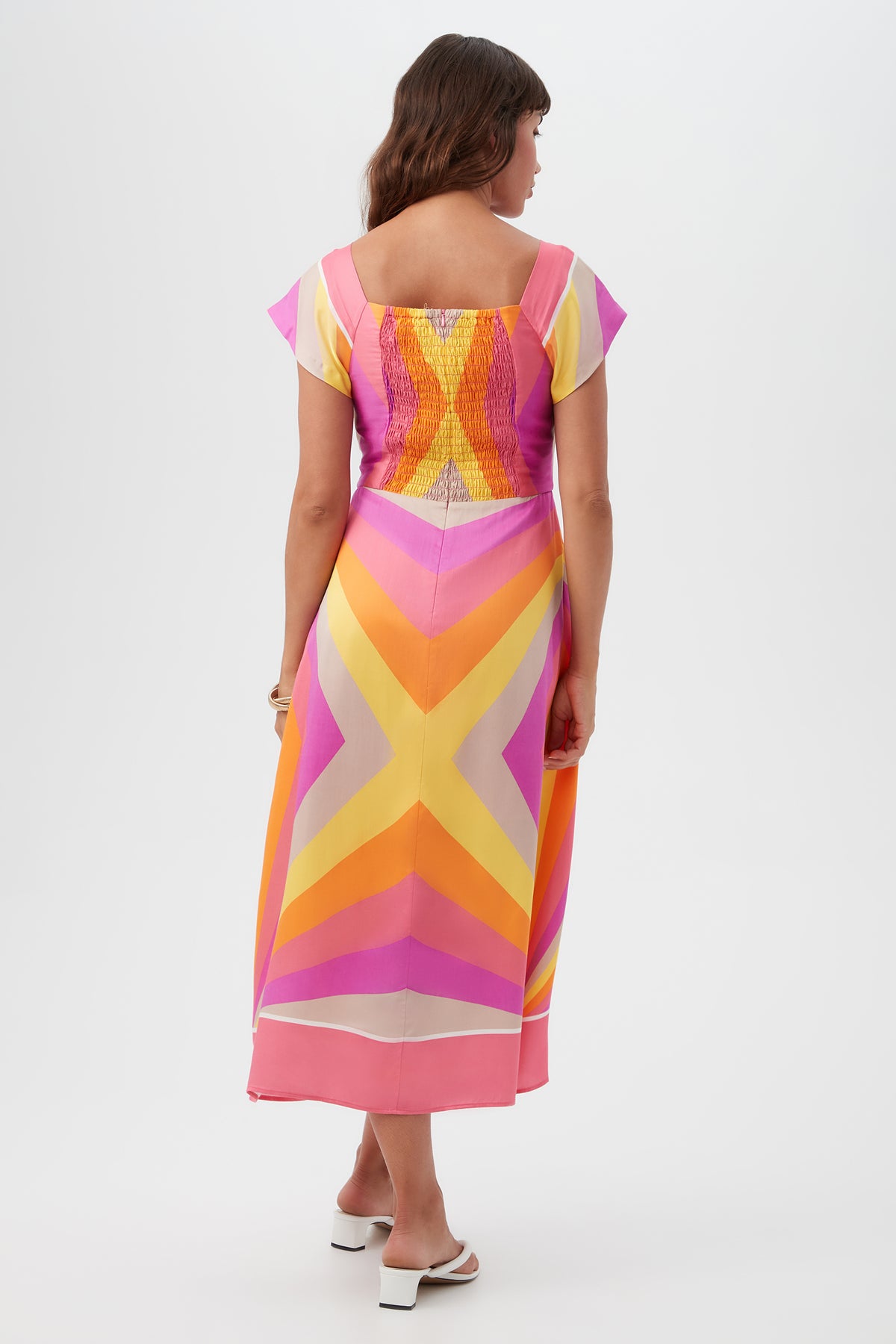 A woman with long brown hair and white heeled sandals faces away, wearing the Après Short-Sleeve Dress featuring pink, yellow, and orange chevron patterns. The photo is set against a plain white background.