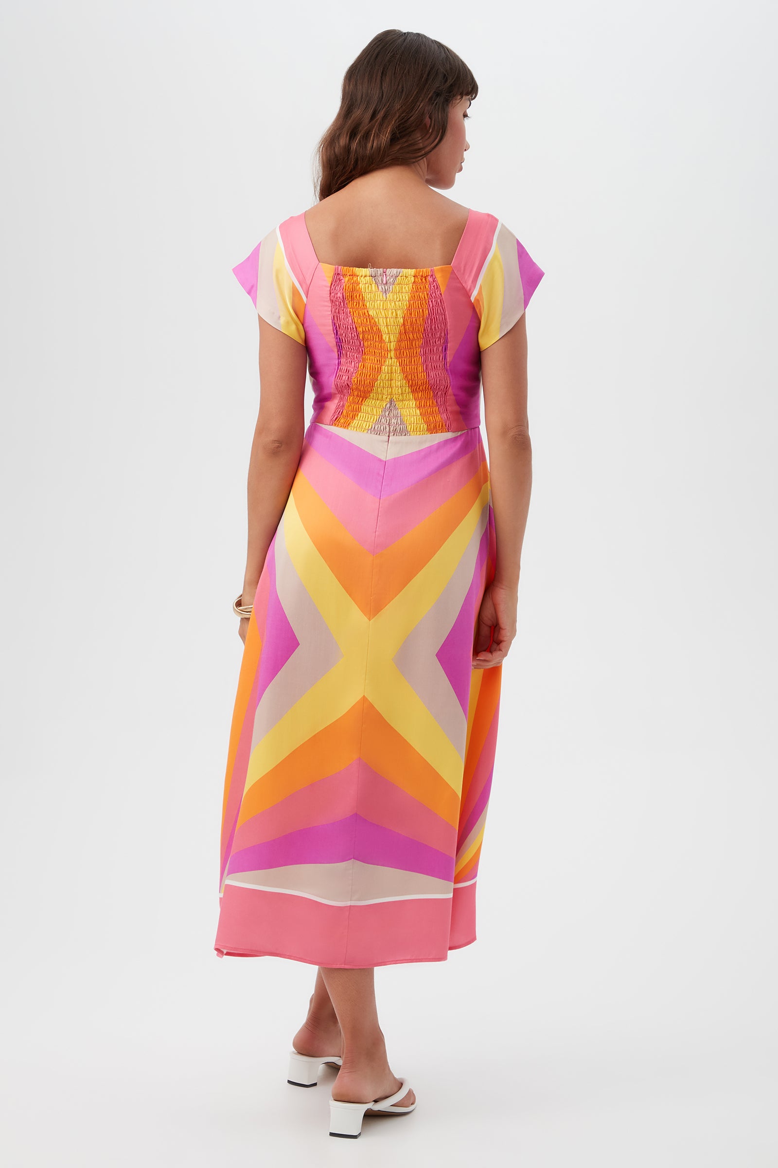 A woman with long brown hair and white heeled sandals faces away, wearing the Après Short-Sleeve Dress featuring pink, yellow, and orange chevron patterns. The photo is set against a plain white background.