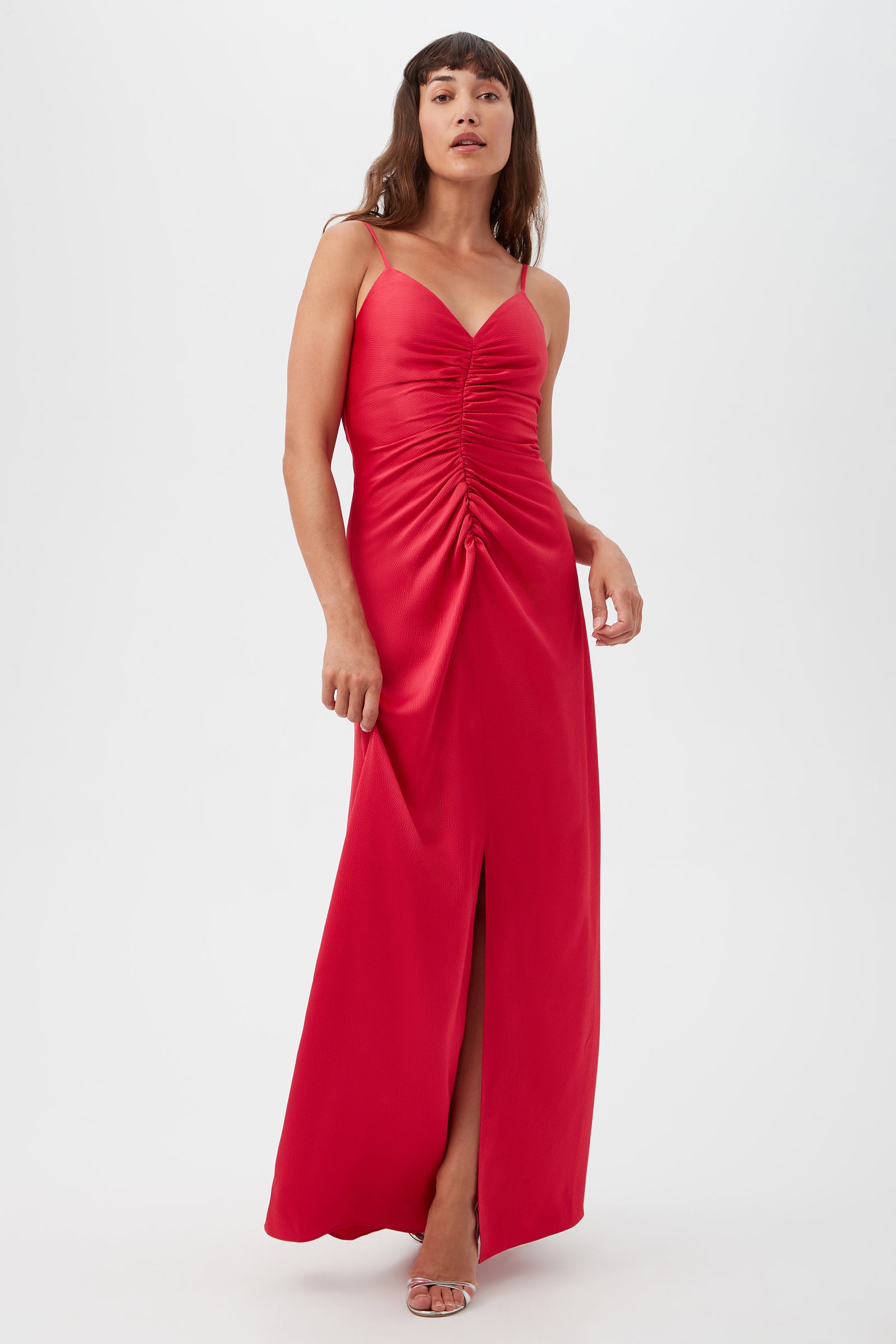 A woman with long brown hair and bangs poses against a white background, wearing the Tulipe Sleeveless Satin Dress in Cannes Coral. The floor-length dress features a ruched front, thigh-high slit, and is styled with open-toe heels.