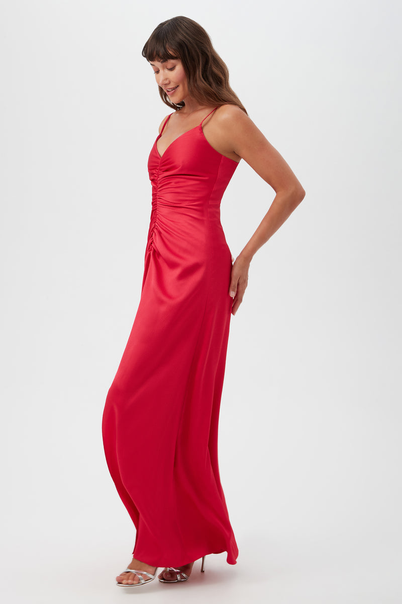 A woman stands sideways, looking down and smiling, in the Tulipe Sleeveless Satin Dress—a long red dress with spaghetti straps and front ruching. She has wavy brown hair and wears silver high-heeled sandals.