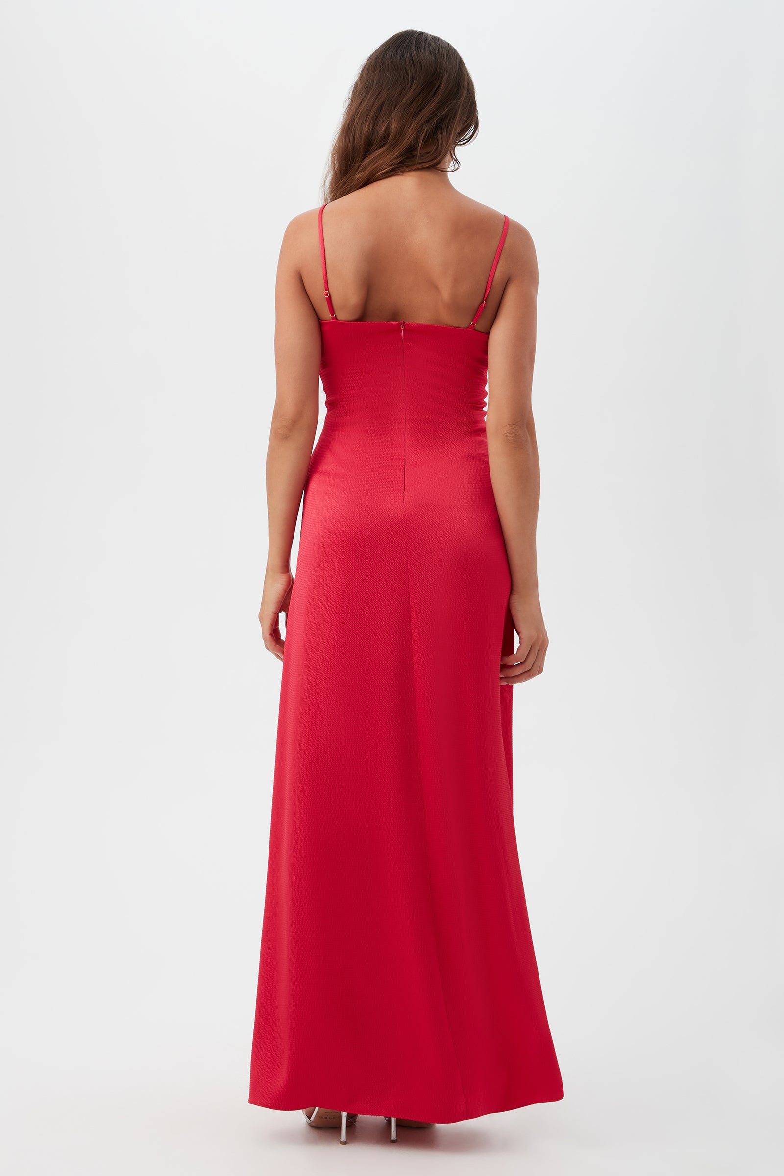 A woman with long hair stands facing away, wearing the Tulipe Sleeveless Satin Dress—a floor-length, red satin gown with thin straps—against a plain white background.