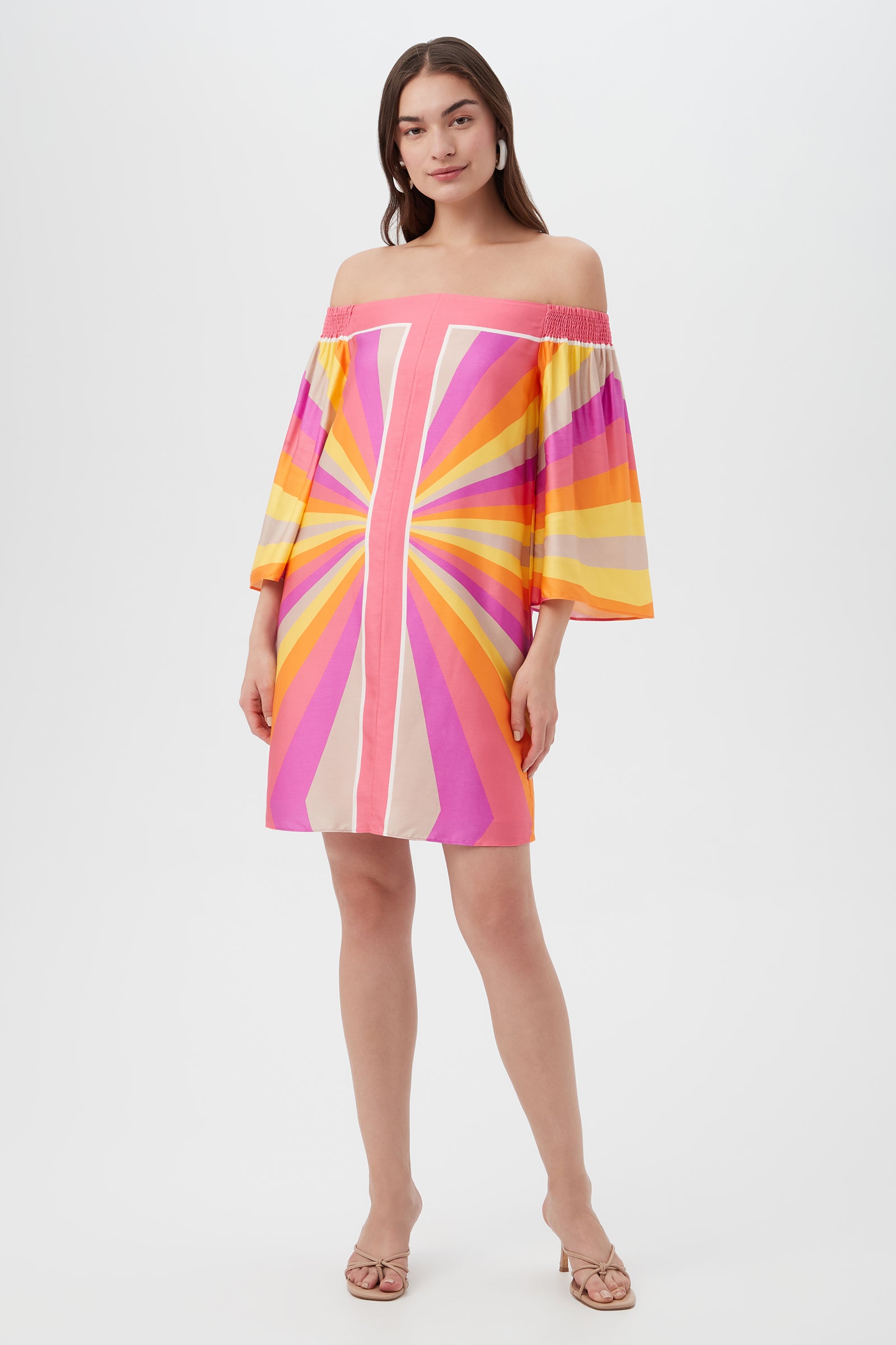 A woman with long brown hair models the Amaris 3 Off-Shoulder Dress in Multi, featuring flared sleeves and a colorful sunburst pattern. She pairs it with sandals and poses against a white background.