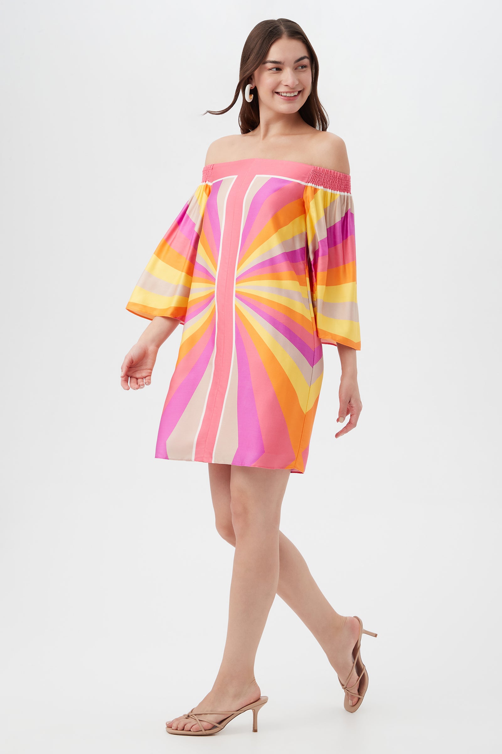 A woman smiles while walking in the Amaris 3 Off-Shoulder Dress, featuring a vibrant geometric pattern in pink, orange, yellow, and purple, paired with beige heeled sandals against a white background.