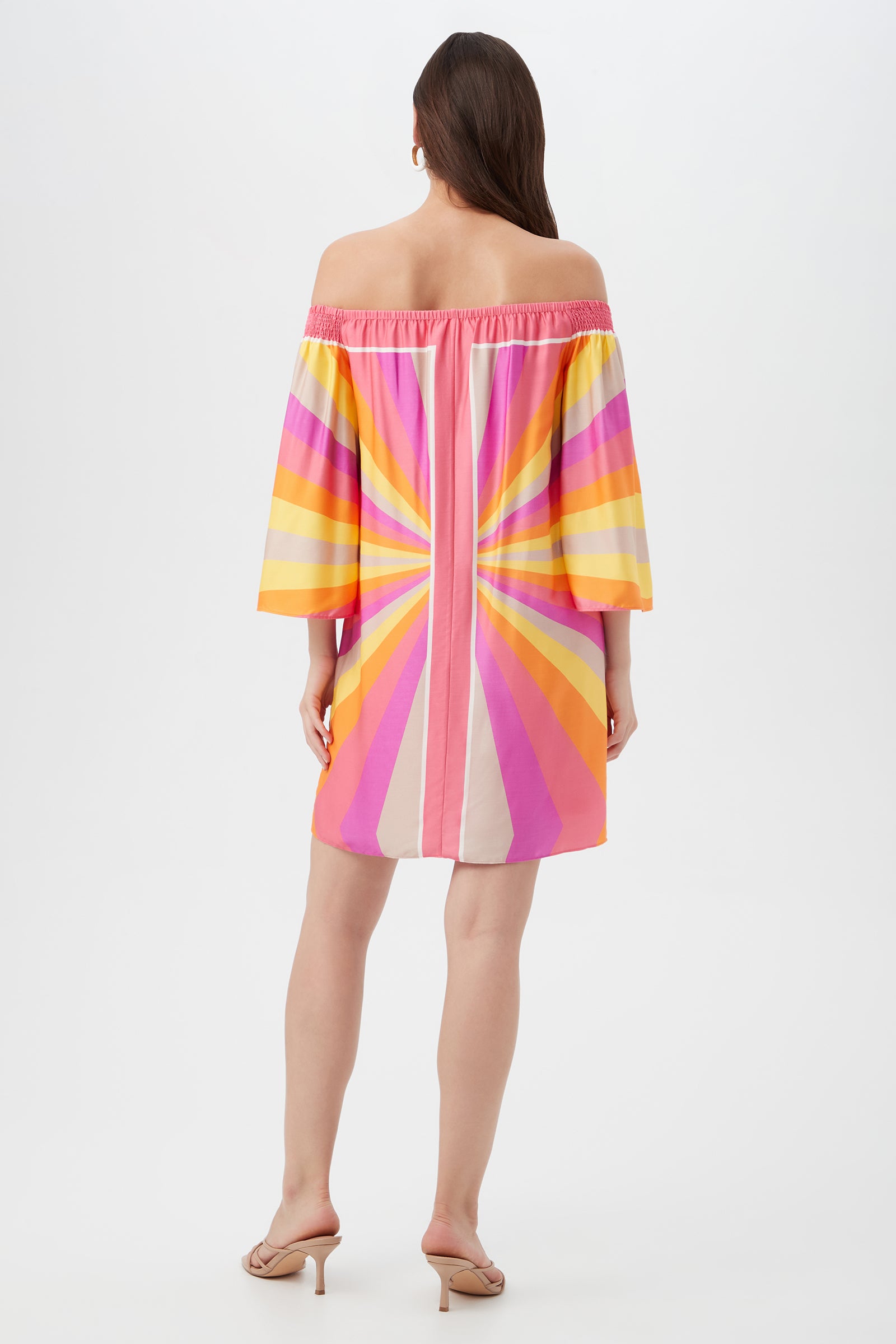 A woman with long brown hair and sandals faces away, wearing the Amaris 3 Off-Shoulder Dress featuring wide sleeves and a bold sunburst pattern in pink, yellow, orange, and peach.