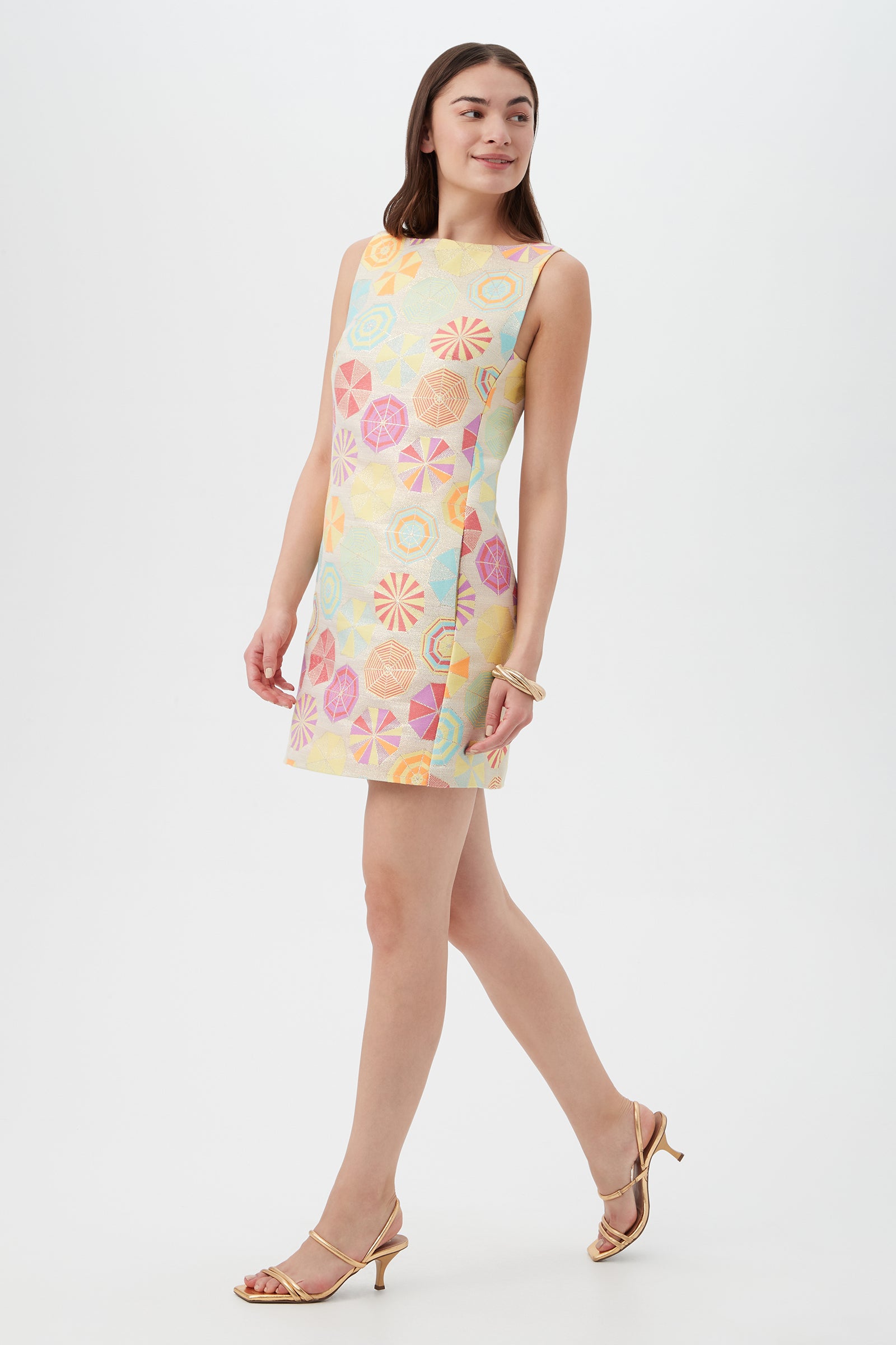A woman wears the TRINA TURK Jalane Sleeveless Dress in Parapluie Jacquard, pairing it with strappy gold heels as she walks confidently against a white background.