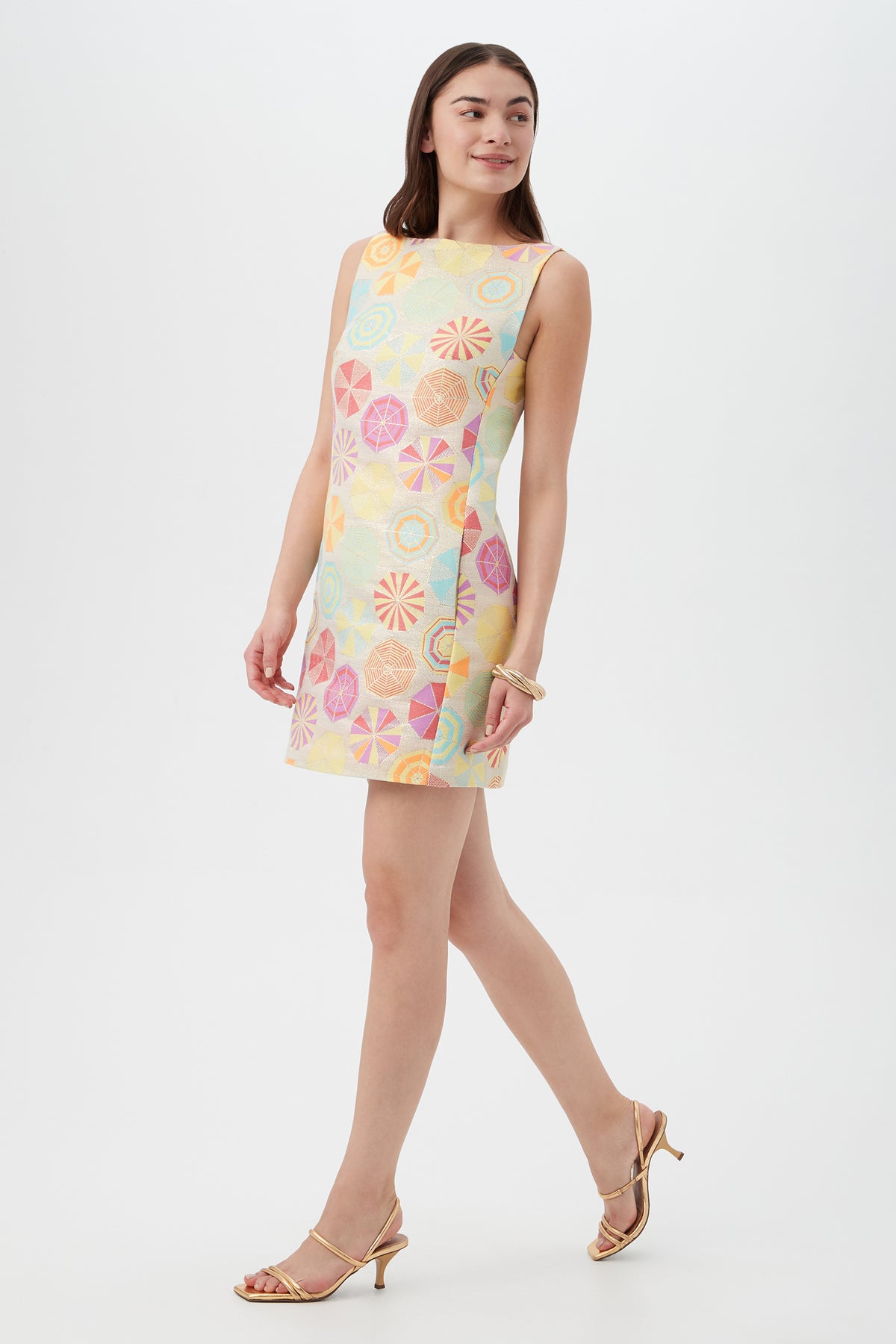 A woman wears the TRINA TURK Jalane Sleeveless Dress in Parapluie Jacquard, pairing it with strappy gold heels as she walks confidently against a white background.