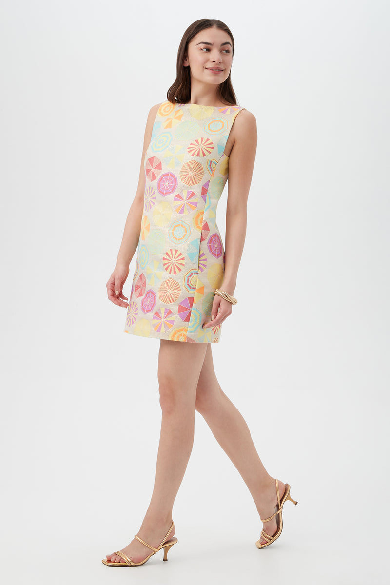 A woman wears the TRINA TURK Jalane Sleeveless Dress in Parapluie Jacquard, pairing it with strappy gold heels as she walks confidently against a white background.