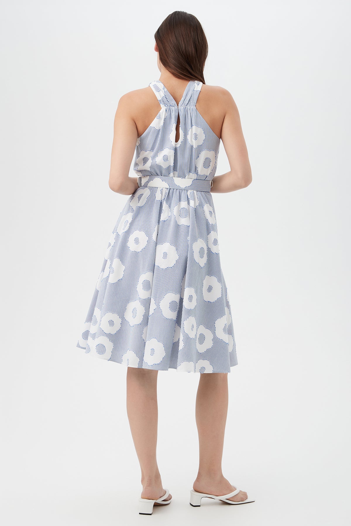 A woman with straight brown hair faces away, wearing the Mardi Sleeveless Dress - Cassis Cotton, a blue and white floral knee-length dress with a halter neckline and open back, paired with white heeled sandals.