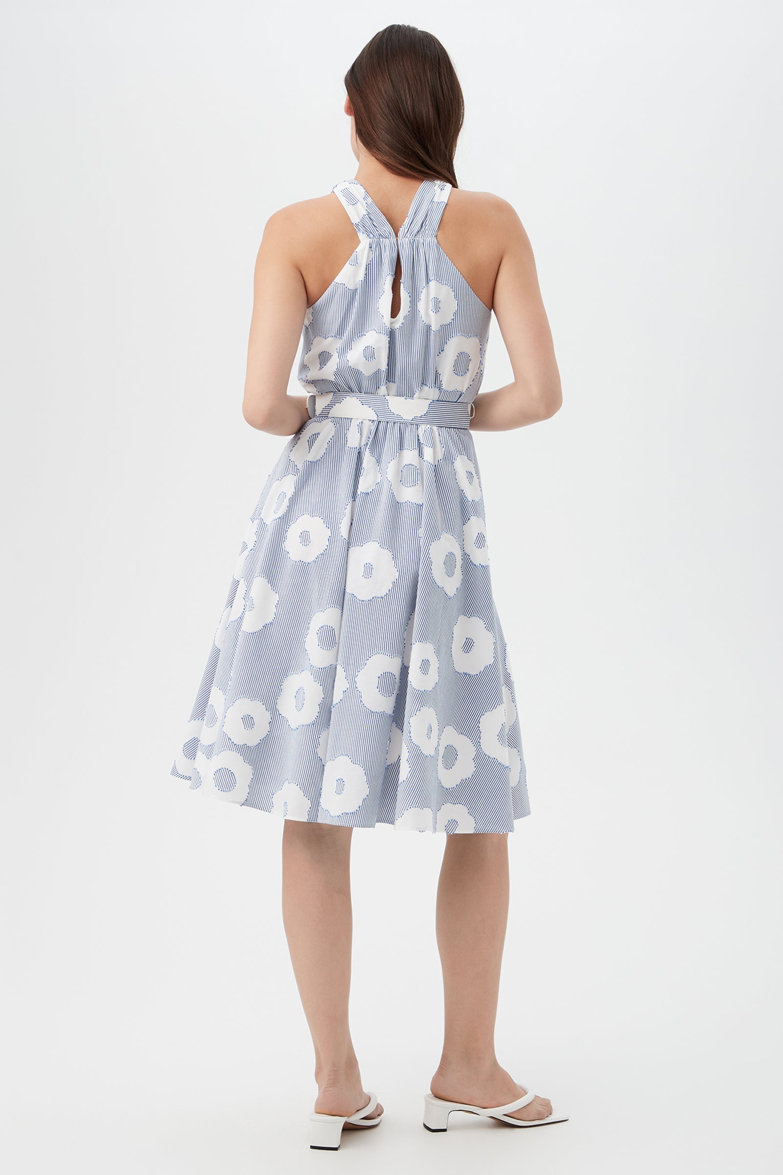 A woman with straight brown hair faces away, wearing the Mardi Sleeveless Dress - Cassis Cotton, a blue and white floral knee-length dress with a halter neckline and open back, paired with white heeled sandals.