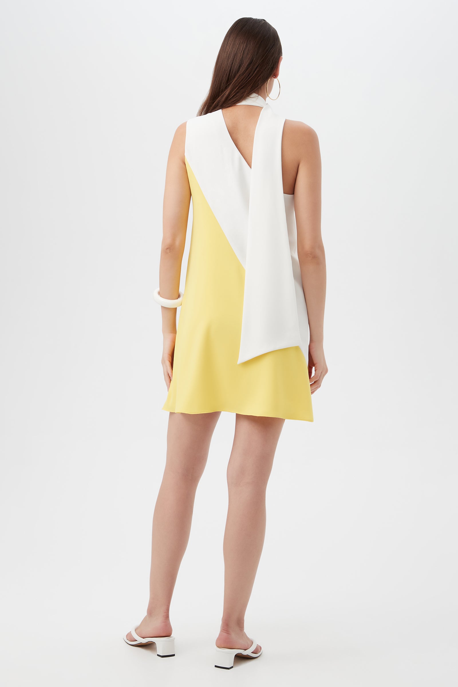 A woman with long brown hair wears the Mielle Sleeveless Color-Block Dress, featuring yellow fabric and a white asymmetrical shoulder panel. She accessorizes with white sandals, large hoop earrings, and a chunky bracelet.