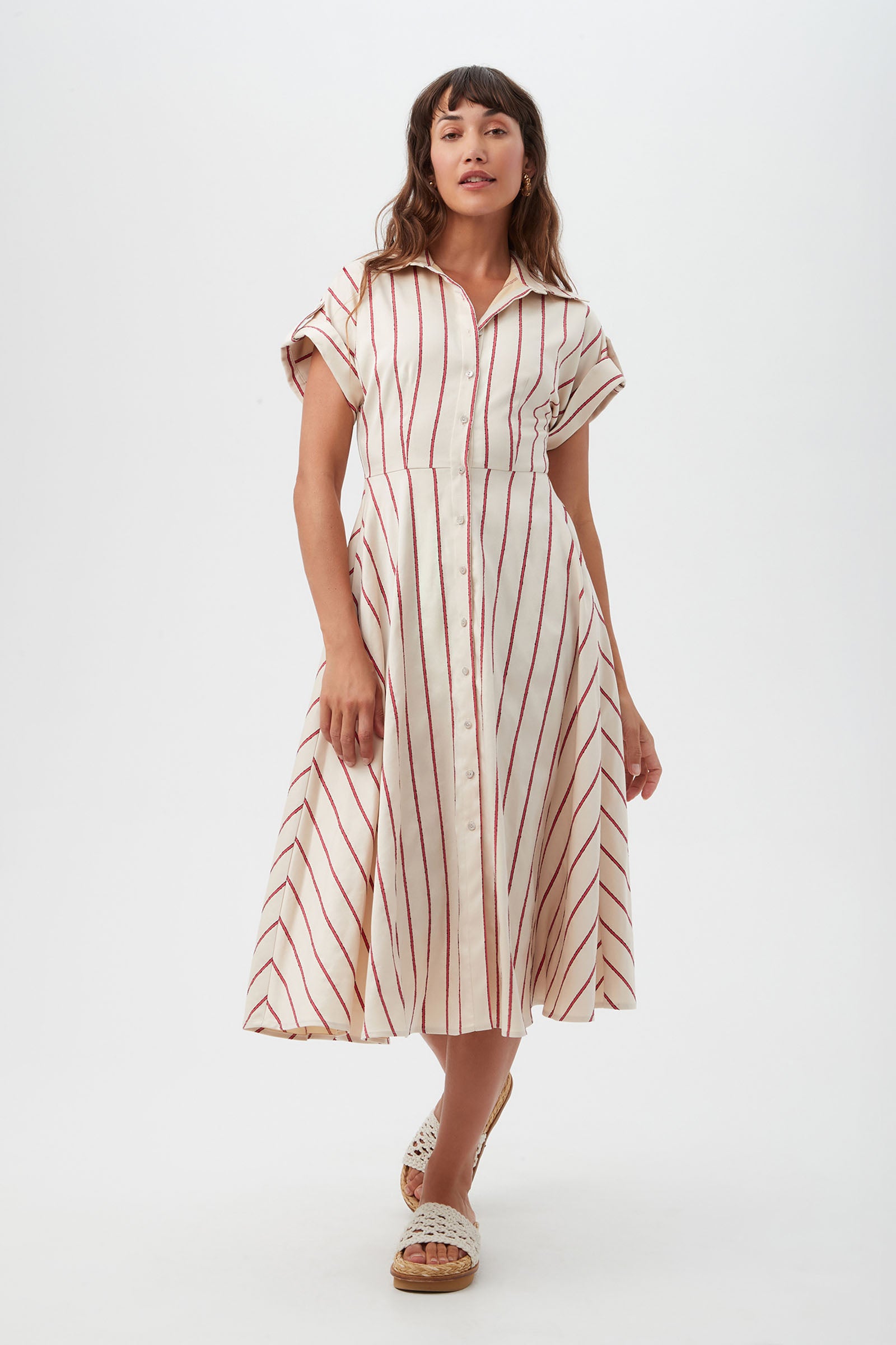 A woman with long, wavy brown hair wears the Lorain Midi Dress - Solaire Stripe, a cream short-sleeve button-up midi dress with red diagonal stripes, paired with white sandals, standing against a plain white background.