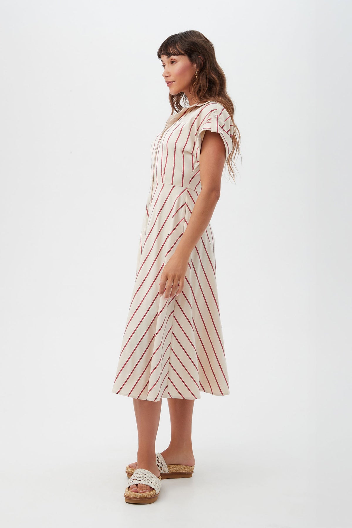 A woman stands sideways against a white background wearing the Lorain Midi Dress - Solaire Stripe, a white, short-sleeved midi dress with red diagonal stripes, paired with tan platform sandals.