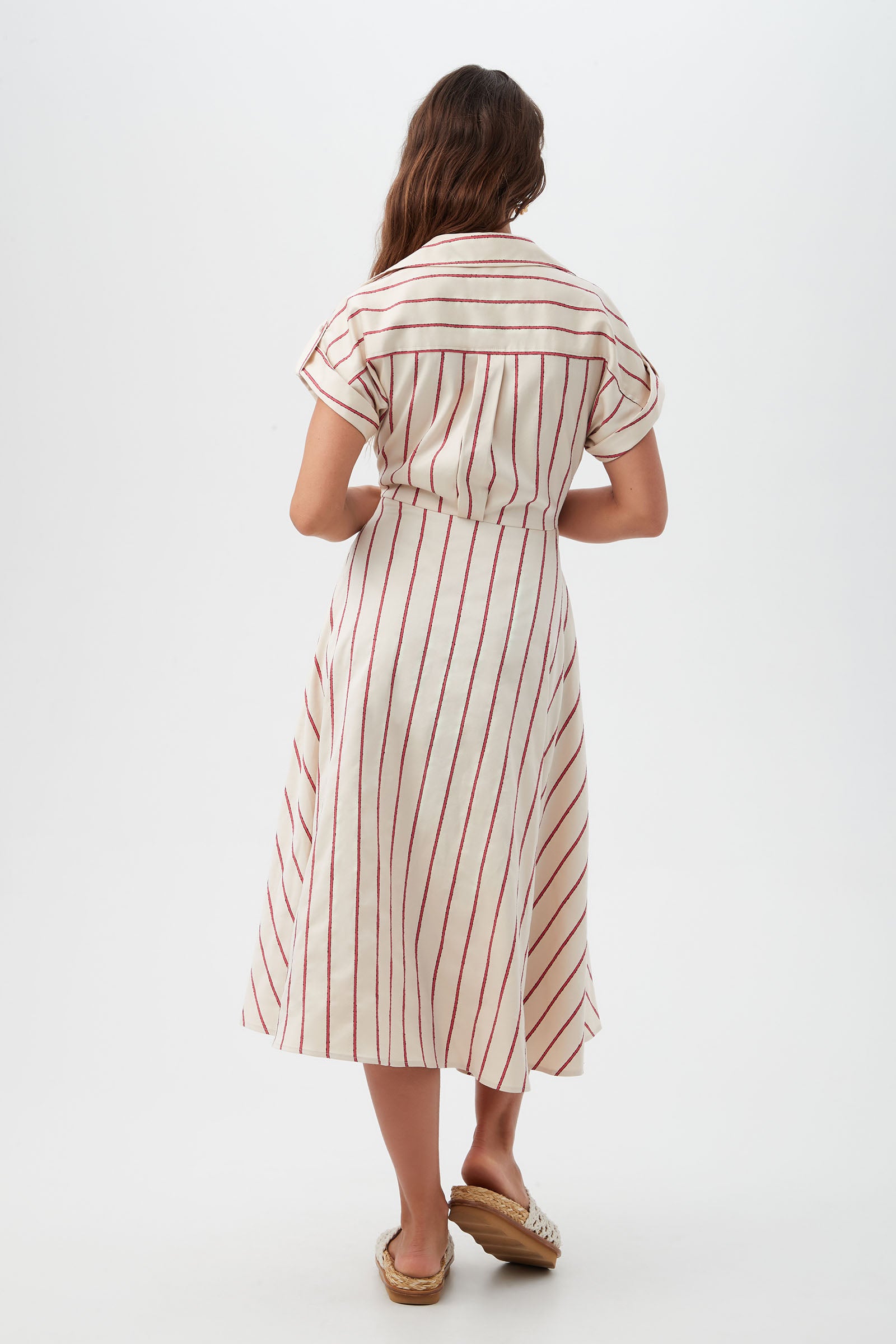 A woman with long brown hair stands facing away, wearing the Lorain Midi Dress - Solaire Stripe, a white short-sleeved, calf-length dress with vertical red stripes and flat woven sandals against a plain light background.