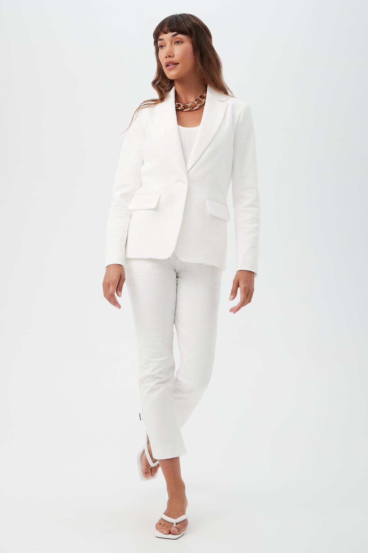 A woman with long brown hair stands against a white background, wearing Saltie 2 Crop Pants with a matching white blazer, white top, white sandals, and a chunky gold chain necklace. She is looking slightly to the side.