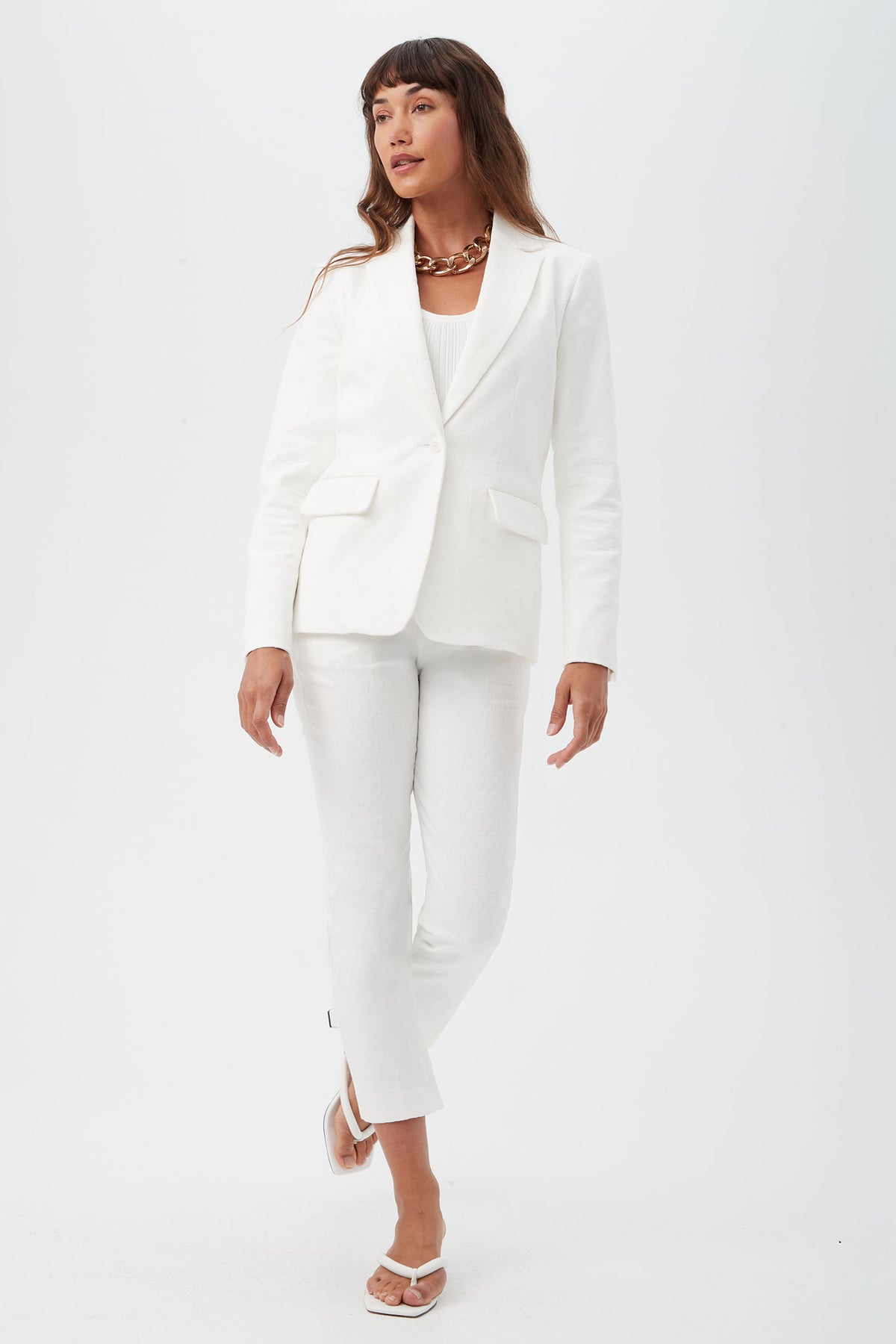 A woman with long brown hair stands against a white background, wearing Pop Slim-Leg Stretch Pants, a white blazer, white top, white sandals, and a chunky necklace. She looks slightly to the side.