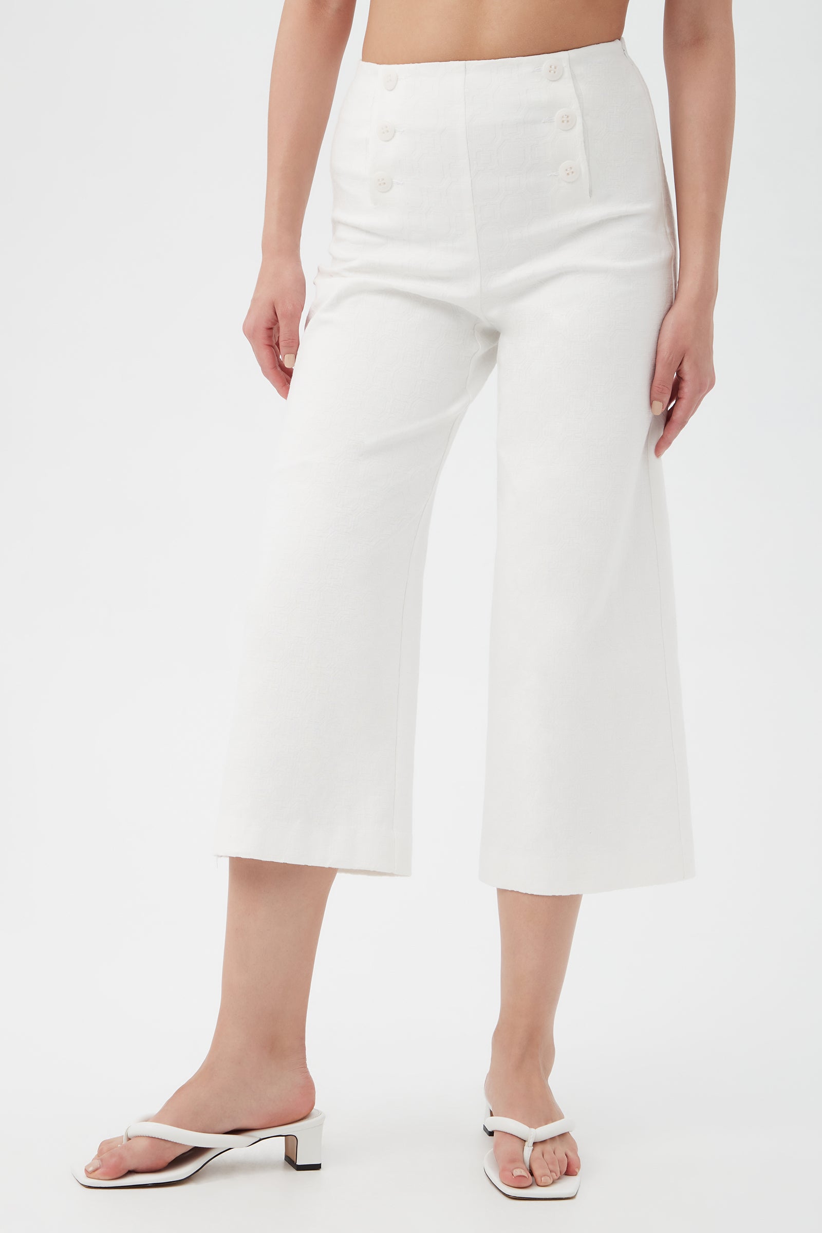 A woman models the Saltie 2 Crop Pants—high-waisted, wide-leg white pants with front button details—paired with white open-toe heeled sandals against a plain white background.