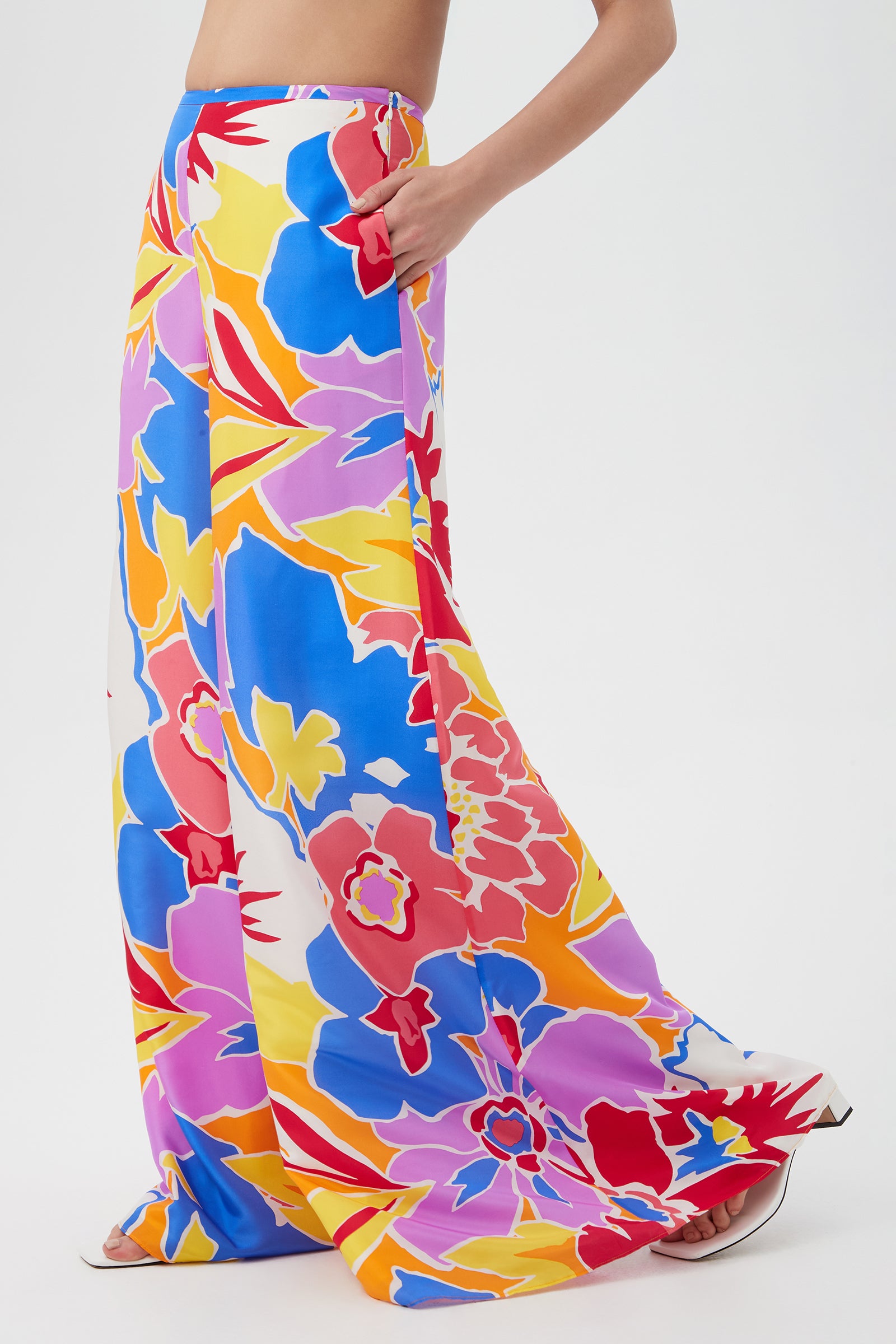 A person wears the Jack Wide-Leg Pant - Soie Paradis, featuring a bold floral print in red, blue, yellow, purple, and white, while standing barefoot against a plain white background.