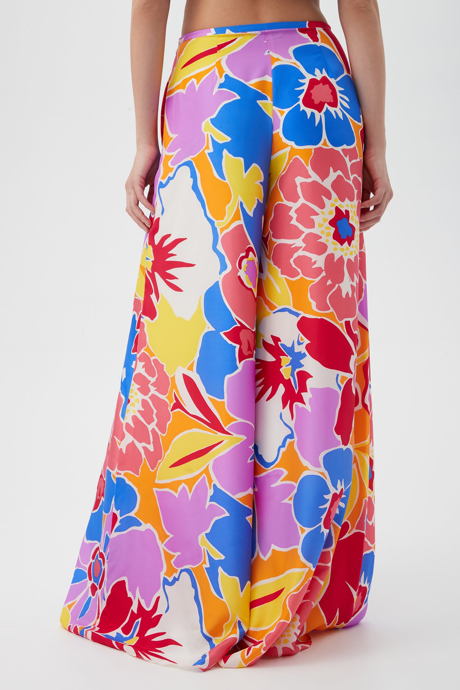 A person wearing the Jack Wide-Leg Pant - Soie Paradis, featuring a vibrant floral pattern in red, yellow, blue, orange, purple, and pink. Photo taken from behind against a plain white background.