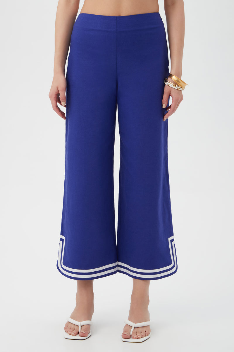 A person wearing Abbey Wide-Leg Crop Pants in cobalt blue with white double stripe trim at the hem, styled with white heeled sandals and gold bangles, stands against a plain white background.