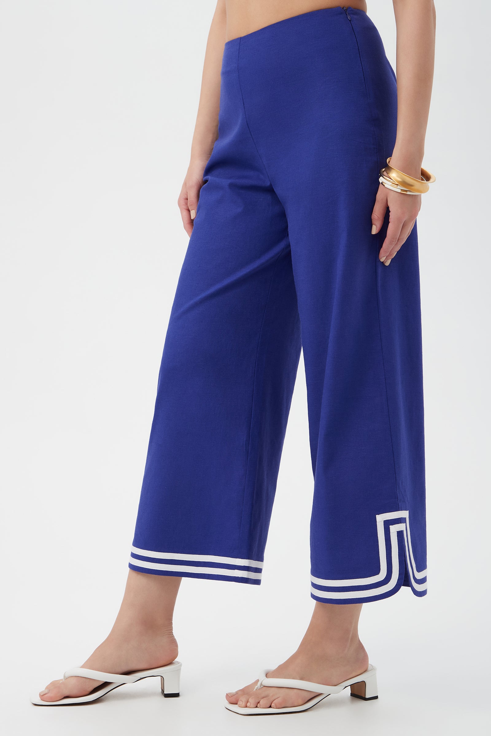 Someone wears the Abbey Wide-Leg Crop Pants in royal blue with white geometric trim at the hem, paired with white heeled sandals and gold bangles, standing against a plain white background.