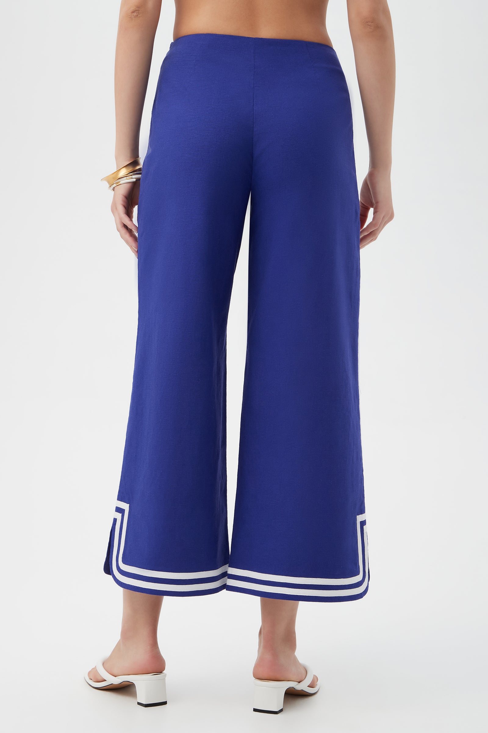 Captured from behind against a white background, a person wears Abbey Wide-Leg Crop Pants in Cobalt Cove with white trim at the hems, paired with white heeled sandals and gold bangles.
