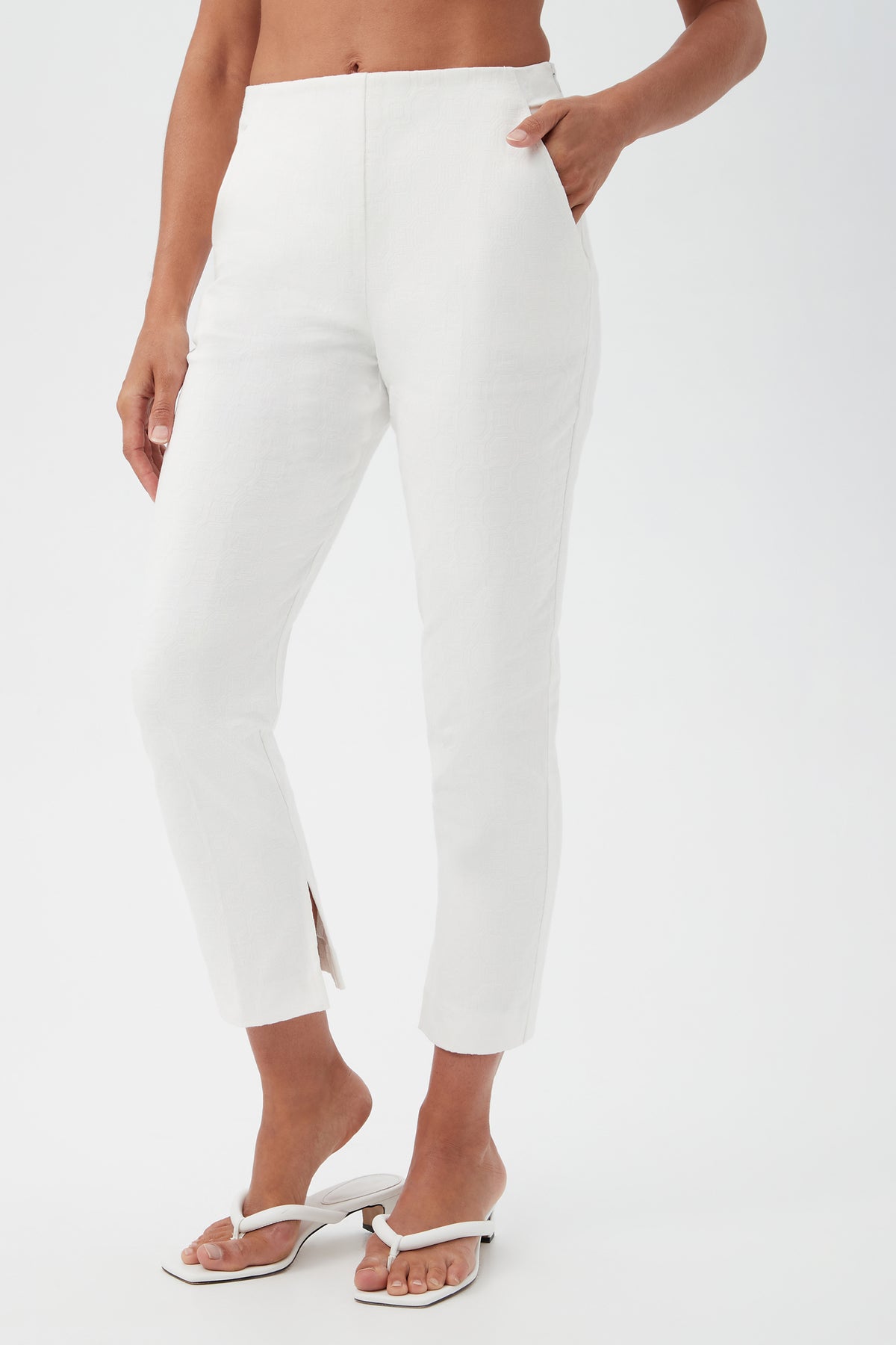 A person wearing Pop Slim-Leg Stretch Pants—white, ankle-length with a small slit at the hem—and white heeled sandals, posing with one hand in their pocket against a plain background.