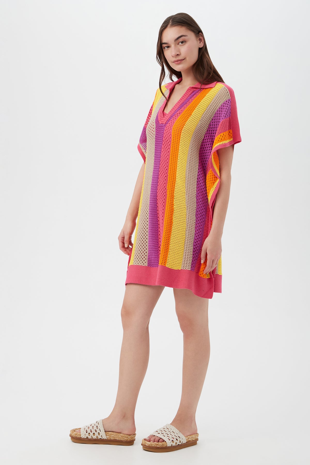 A woman with light skin and straight brown hair wears the Candela Dress, a loose, short-sleeved V-neck knit featuring vertical pink, purple, orange, and yellow stripes. She pairs it with white woven slide sandals against a plain white background.