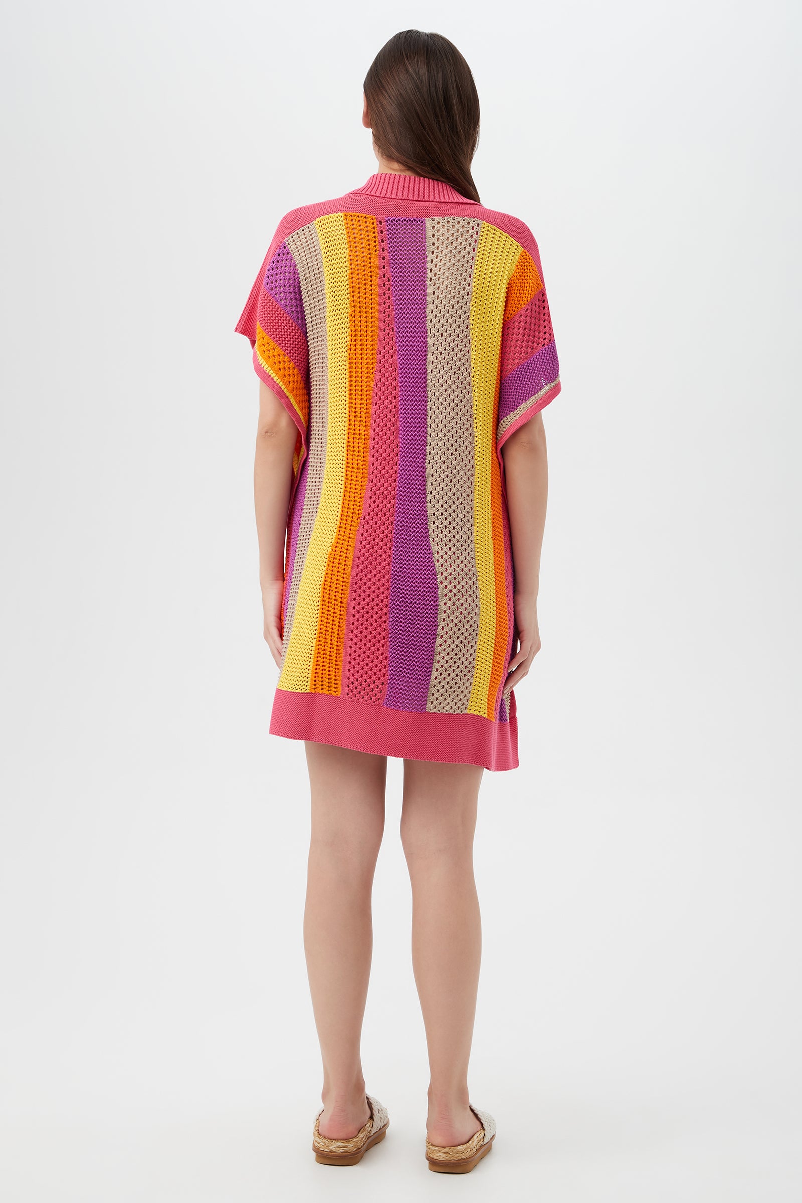 A woman with long brown hair stands facing away in the Candela Dress— a short-sleeved knit dress with vertical pink, orange, yellow, purple, and beige stripes. She wears sandals against a plain white background.