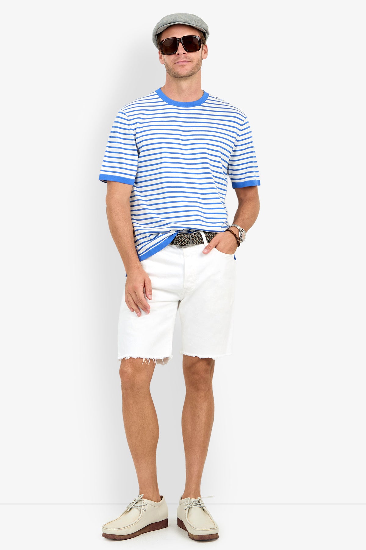 A man wearing a gray flat cap, sunglasses, the Giovanni Stripe Short-Sleeve Pullover, white frayed shorts, a belt, and beige shoes stands against a plain background.