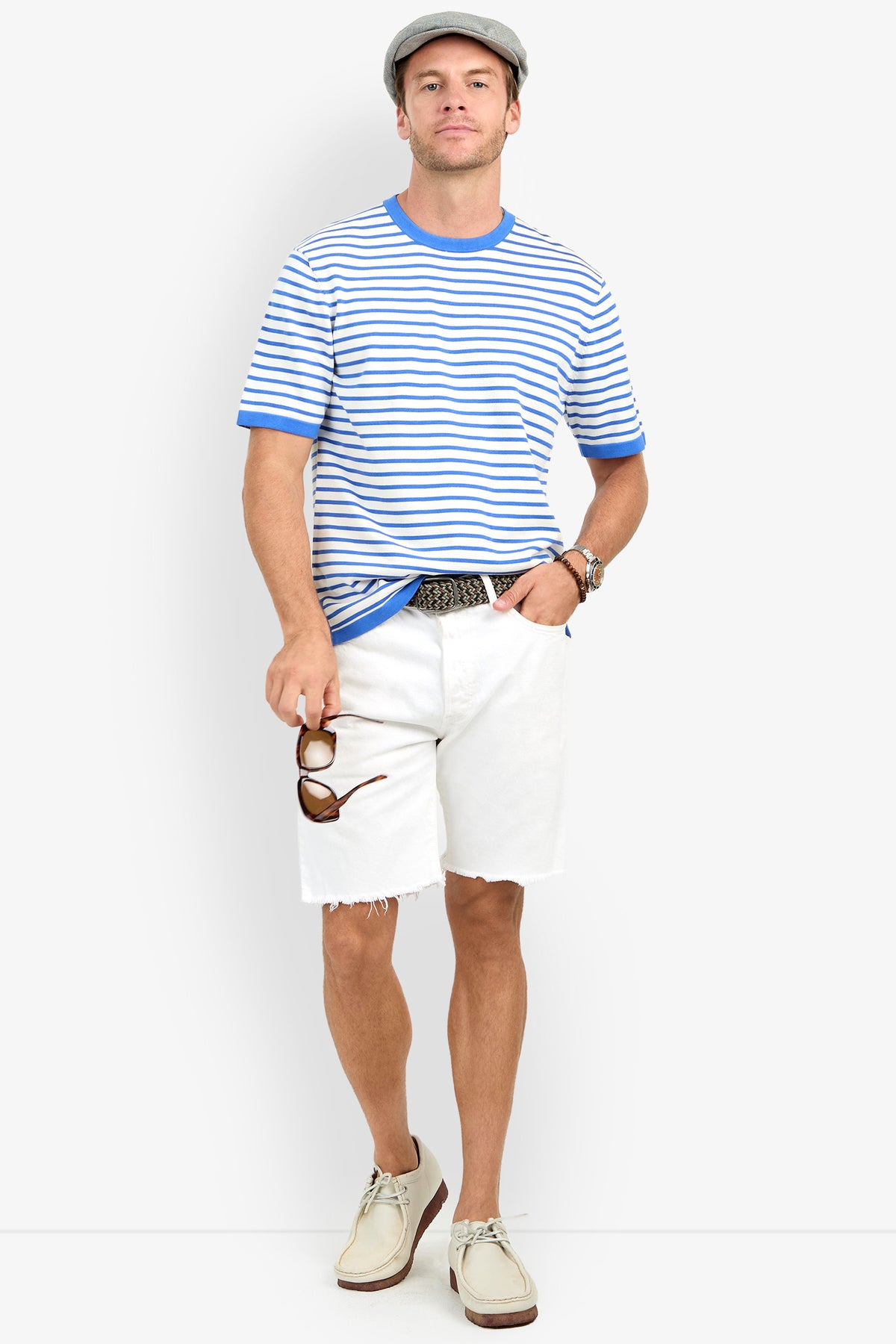 Man in a gray flat cap, Giovanni Stripe Short-Sleeve Pullover, white shorts, beige shoes, and holding sunglasses stands relaxed against a white background.