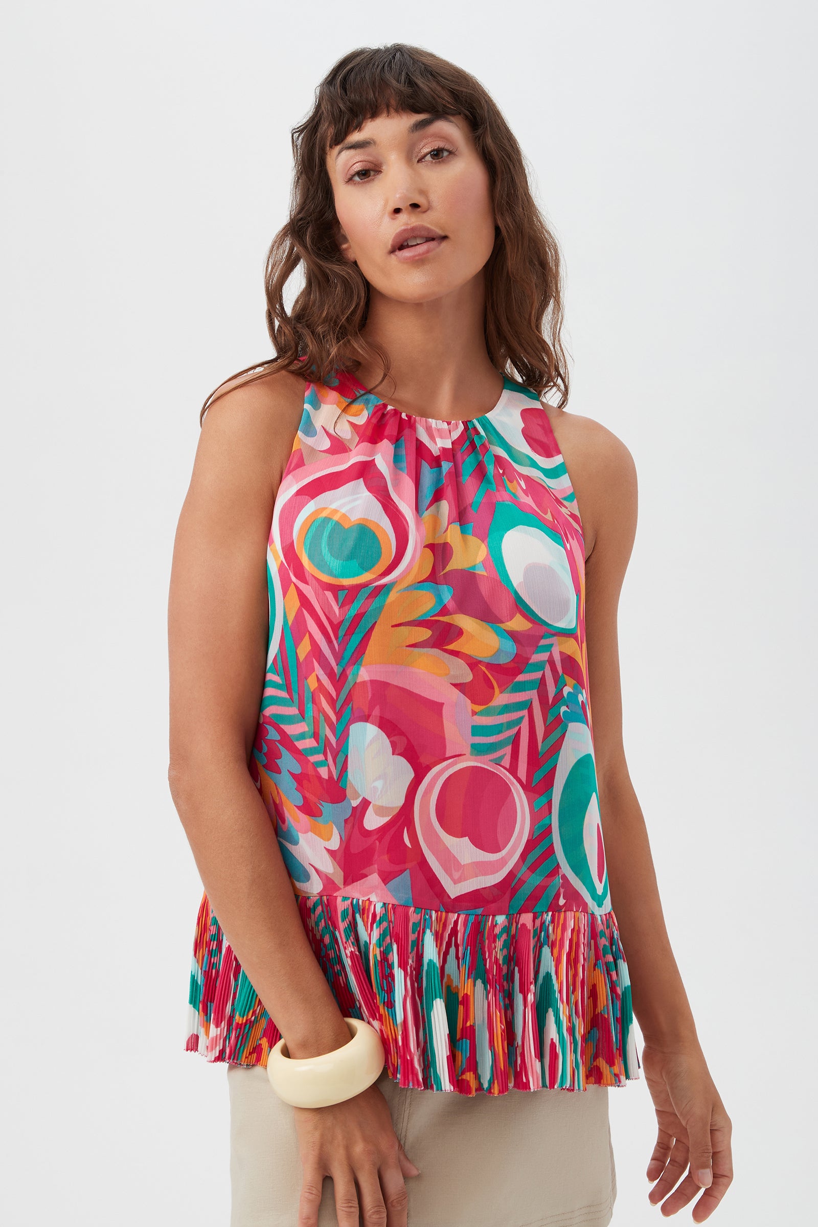 A woman with wavy brown hair wears the Nimes Sleeveless Top in Dramont, featuring a colorful pattern and ruffled hem, paired with a chunky cream bangle against a plain light background.