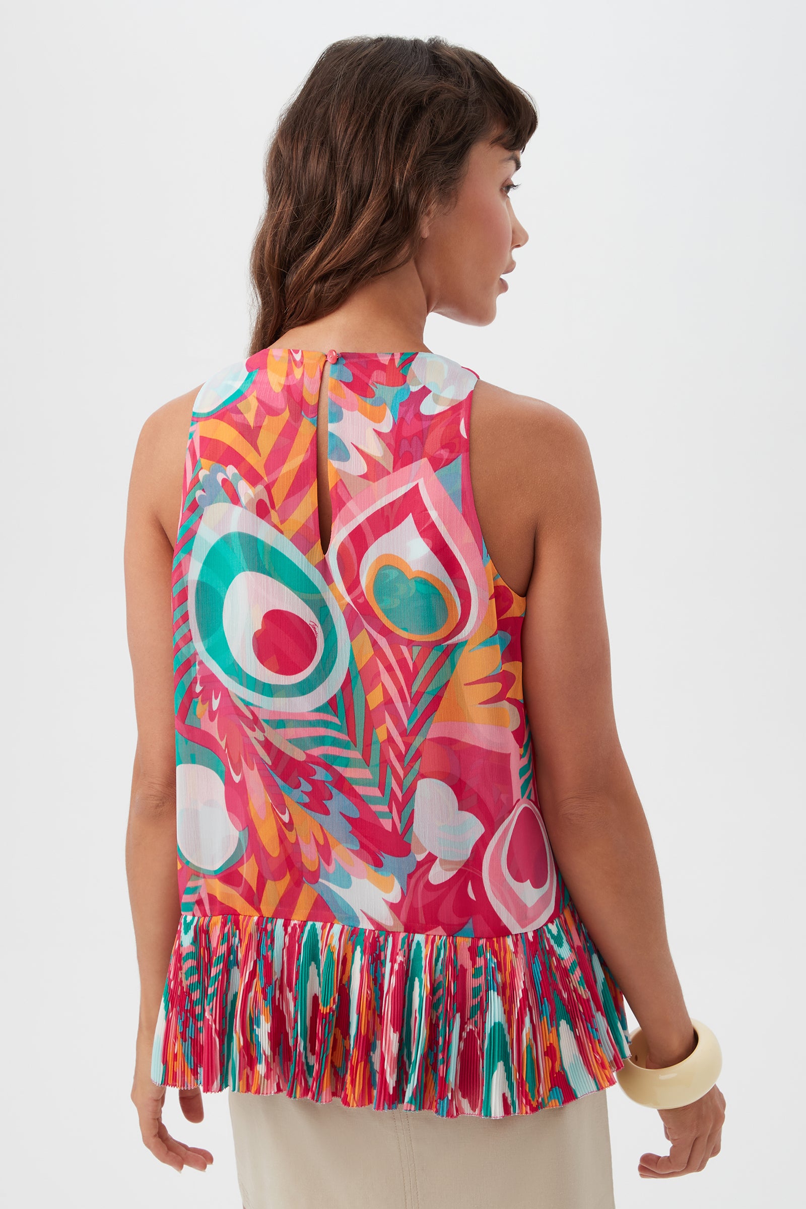 A woman with wavy brown hair and a chunky white bracelet wears the Nimes Sleeveless Top - Dramont, featuring bold, abstract peacock feather patterns and a pleated hem. She faces away against a plain white background.