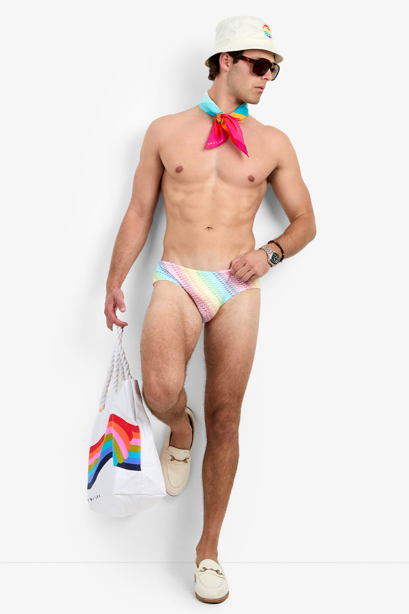 A man wearing the Bondi Swim Brief - Pride, a white bucket hat, sunglasses, and a colorful neck scarf poses with a rainbow tote bag. He also sports white loafers and a wristwatch.