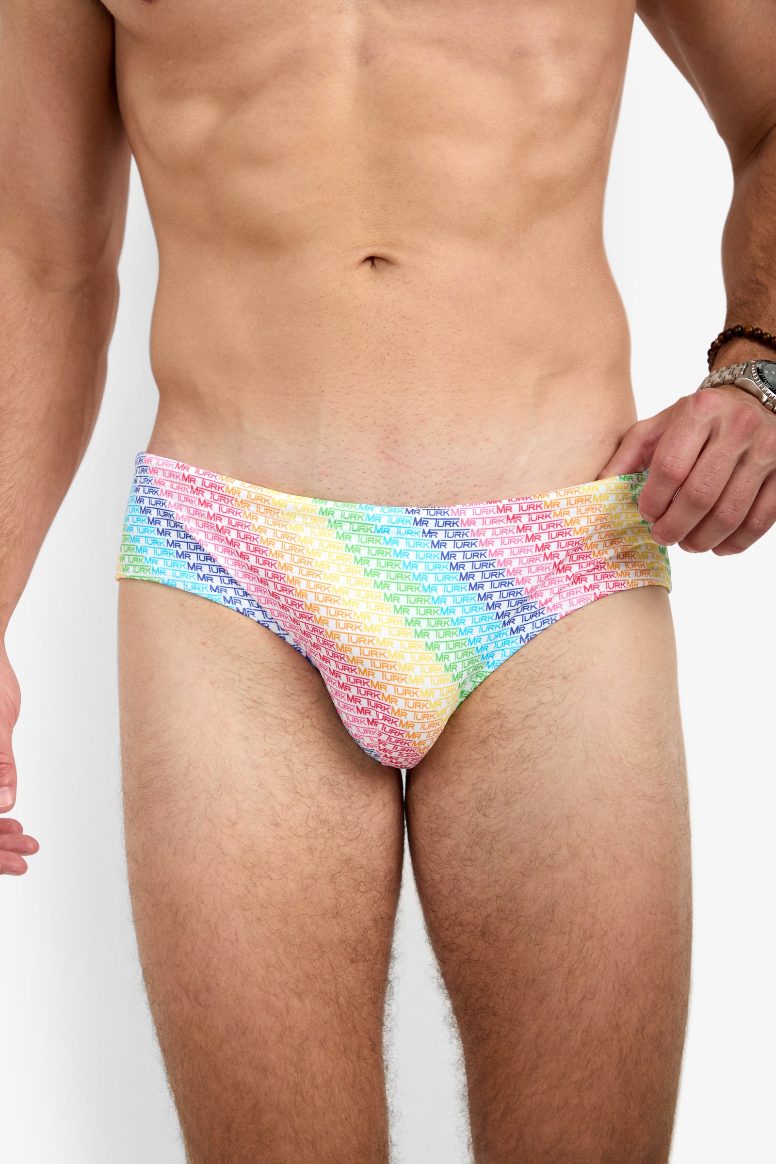 A man models the Bondi Swim Brief - Pride, featuring a rainbow gradient with repeating white text. His torso and upper thighs are visible as he holds the waistband with one hand.