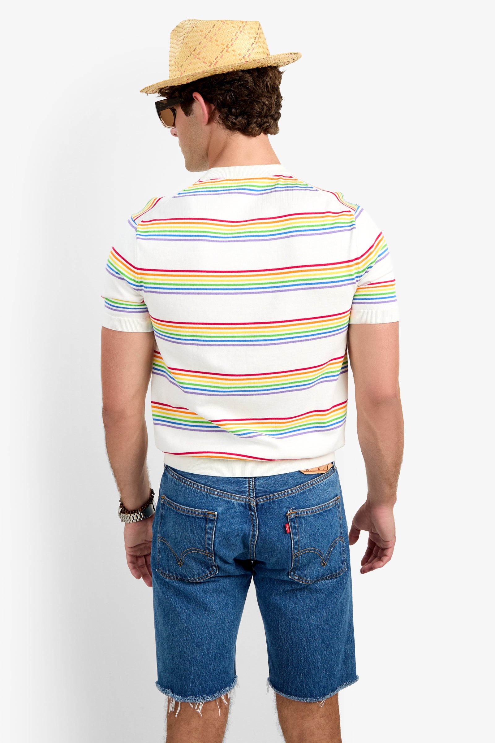 A man stands facing away, wearing a straw hat, sunglasses, a Carlo Short-Sleeve Crewneck - Pride T-shirt with colorful stripes, blue frayed denim shorts, and a bracelet on his left wrist.