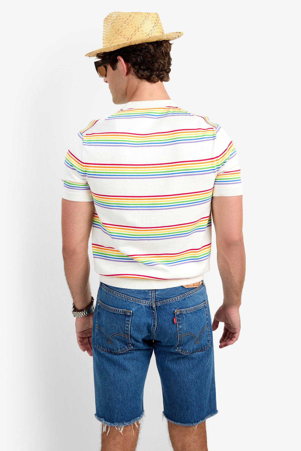 A man stands facing away, wearing a straw hat, sunglasses, a Carlo Short-Sleeve Crewneck - Pride T-shirt with colorful stripes, blue frayed denim shorts, and a bracelet on his left wrist.