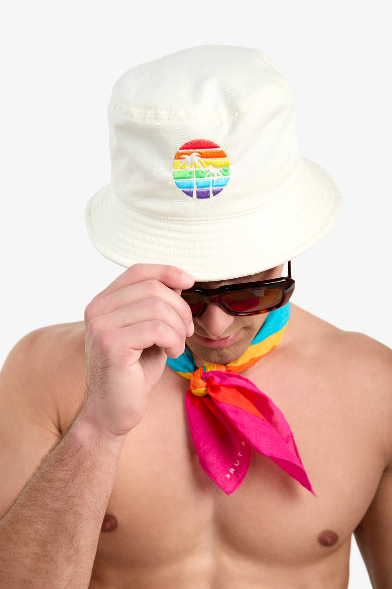 A shirtless person wears the Bucket Hat - Pride featuring a rainbow sunset design, along with dark sunglasses and a colorful blue, yellow, and pink scarf against a plain background.