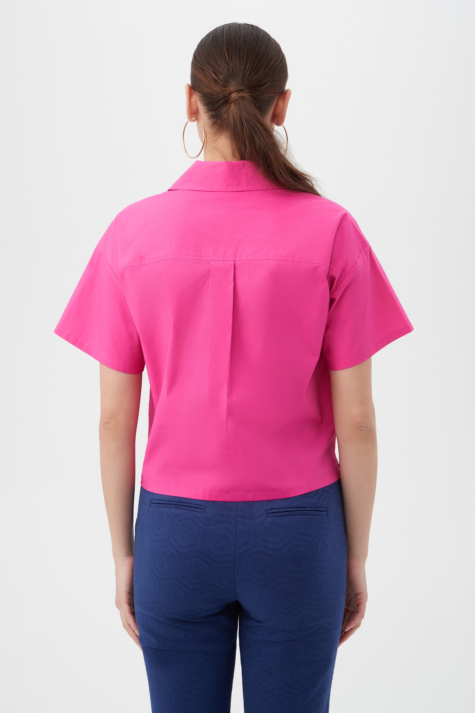 A woman is seen from behind in a Komore Cropped Button-Front Top—bright pink, short-sleeve—paired with blue pants. She has straight brown hair tied back, wears large hoop earrings, and stands against a plain white background.