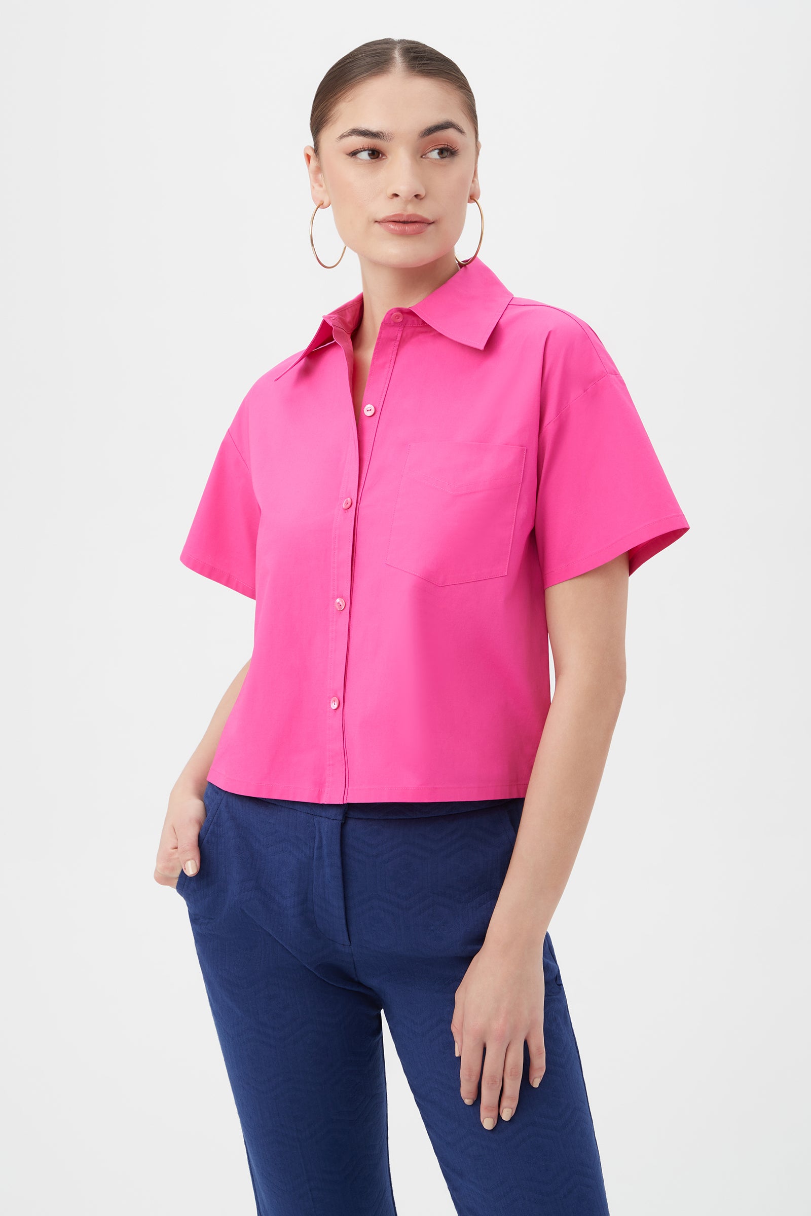 A woman wears the Komore Cropped Button-Front Top in Trina Pink with blue pants. The short-sleeve shirt features a collar and chest pocket. She has her hair pulled back, large hoop earrings, and stands against a plain white background.