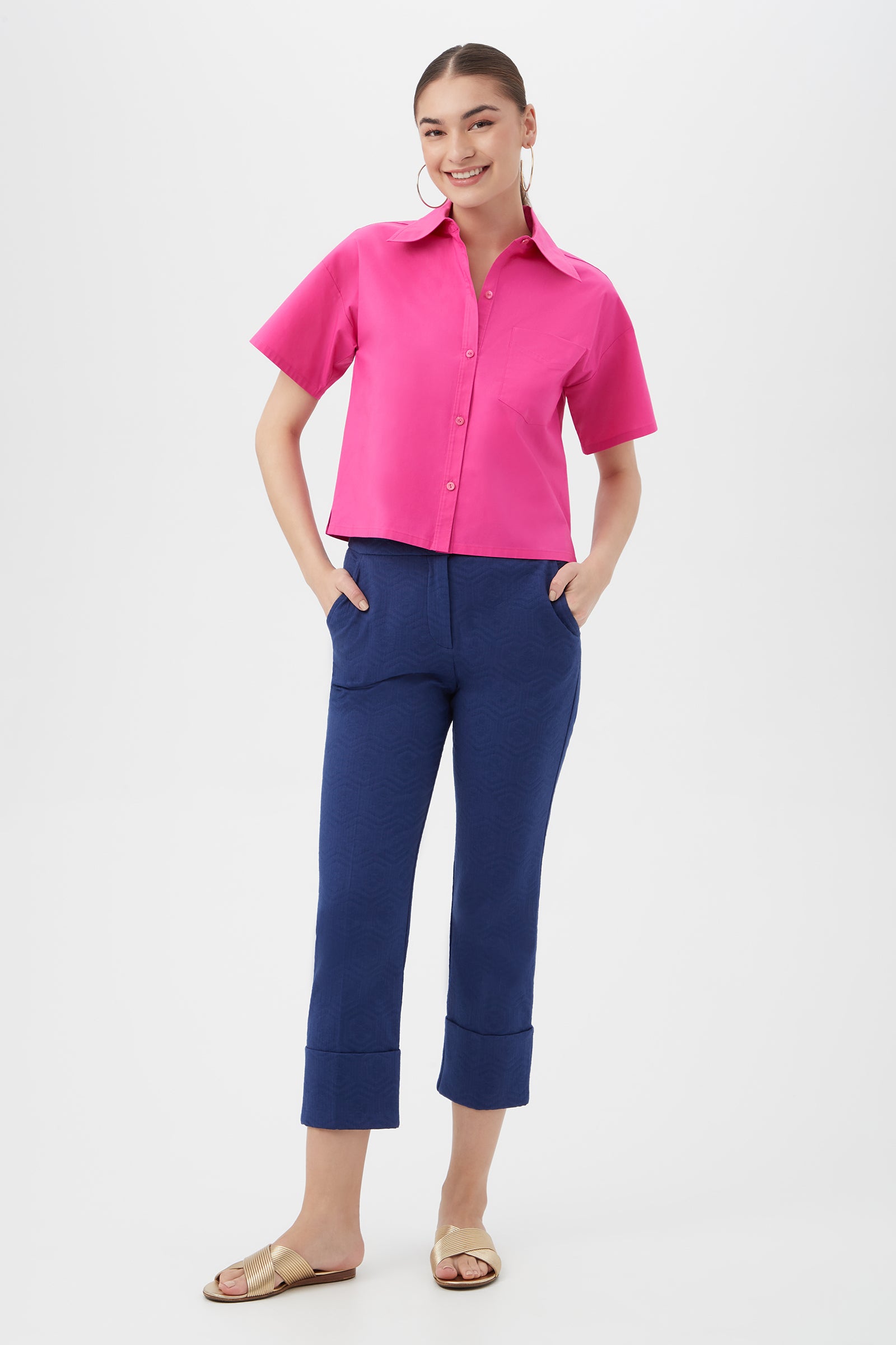 A woman smiles with her hands in her pockets, wearing the Komore Cropped Button-Front Top in bright pink, blue cropped pants, and beige sandals against a plain white background.