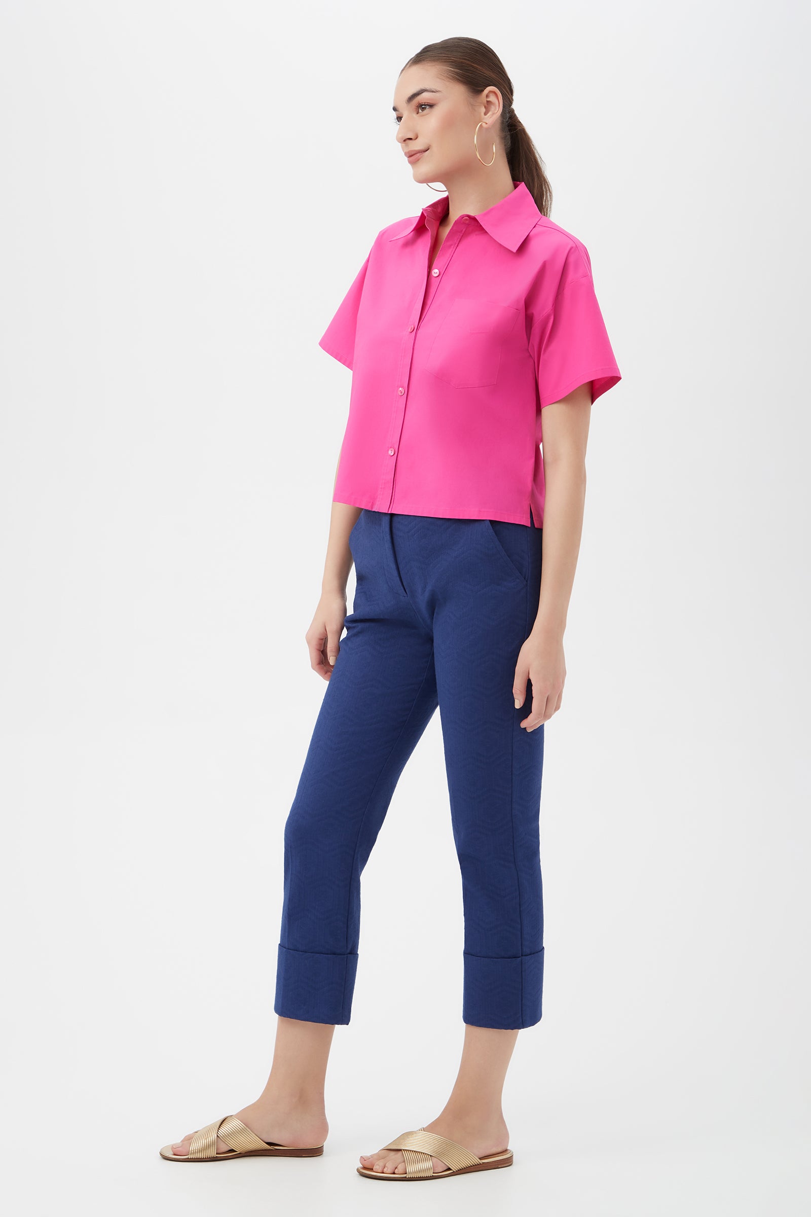 A woman stands against a white background, wearing the Komore Cropped Button-Front Top in bright pink, navy blue cuffed cropped pants, gold hoop earrings, and beige slide sandals.