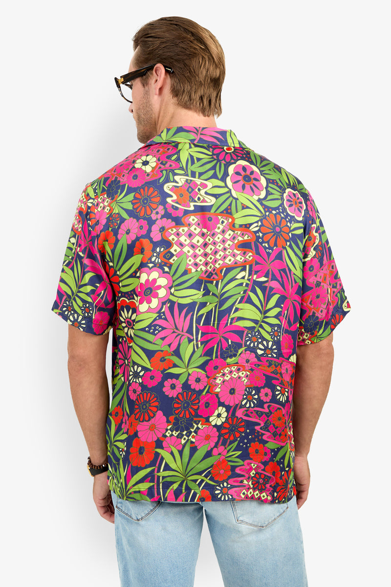 A man wearing glasses and the Felix Camp Shirt, a colorful short-sleeve floral shirt with green, pink, red, and yellow patterns, stands facing away. He also wears light blue jeans.