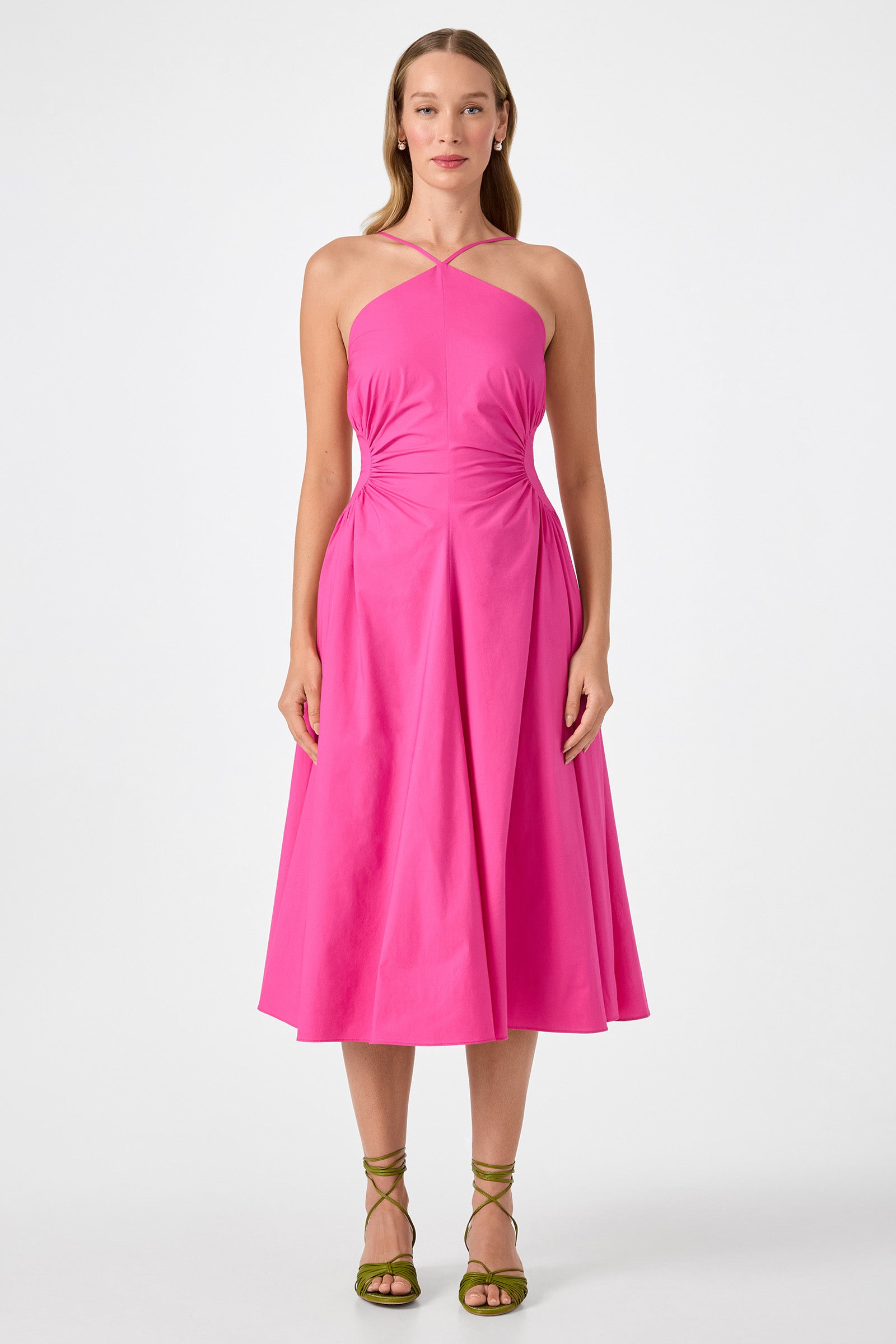 A woman with long hair faces forward, wearing the bright pink Kata Shirred Waist Midi Dress with a halter neck, sleeveless design, and flared skirt. She pairs it with green strappy sandals against a plain white background.