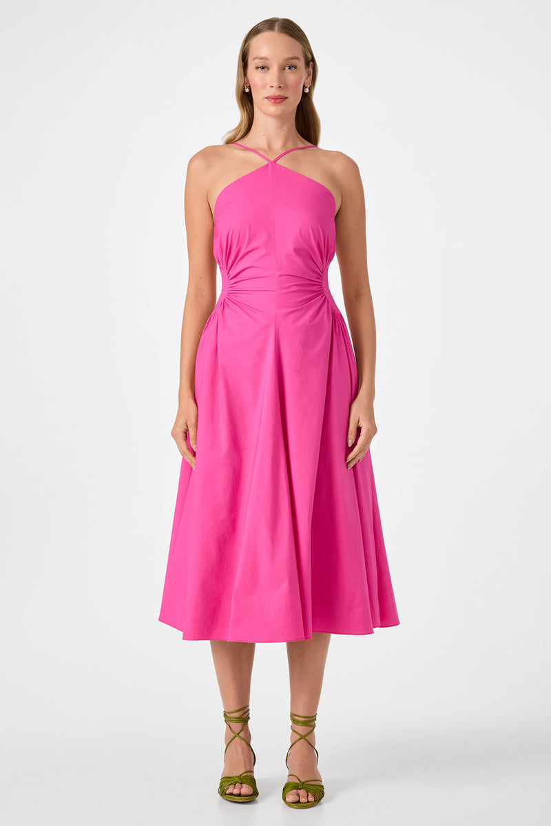 A woman with long hair faces forward, wearing the bright pink Kata Shirred Waist Midi Dress with a halter neck, sleeveless design, and flared skirt. She pairs it with green strappy sandals against a plain white background.