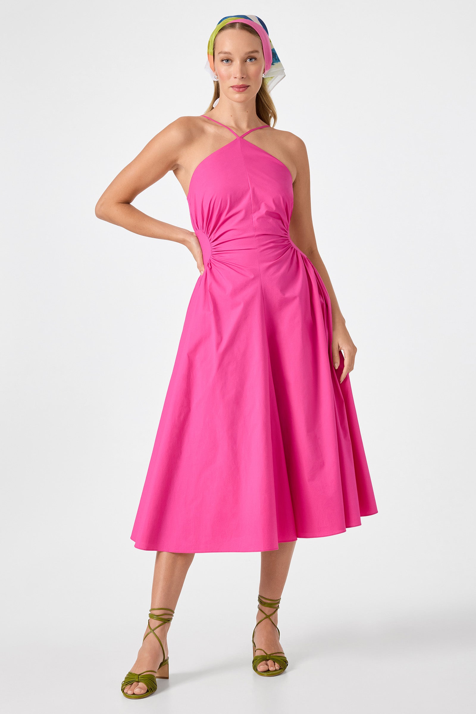 A woman wears the Kata Shirred Waist Midi Dress in Trina Pink with a halter neckline and gathered side detail, styled with a colorful headscarf and green strappy sandals, posing against a plain white background.